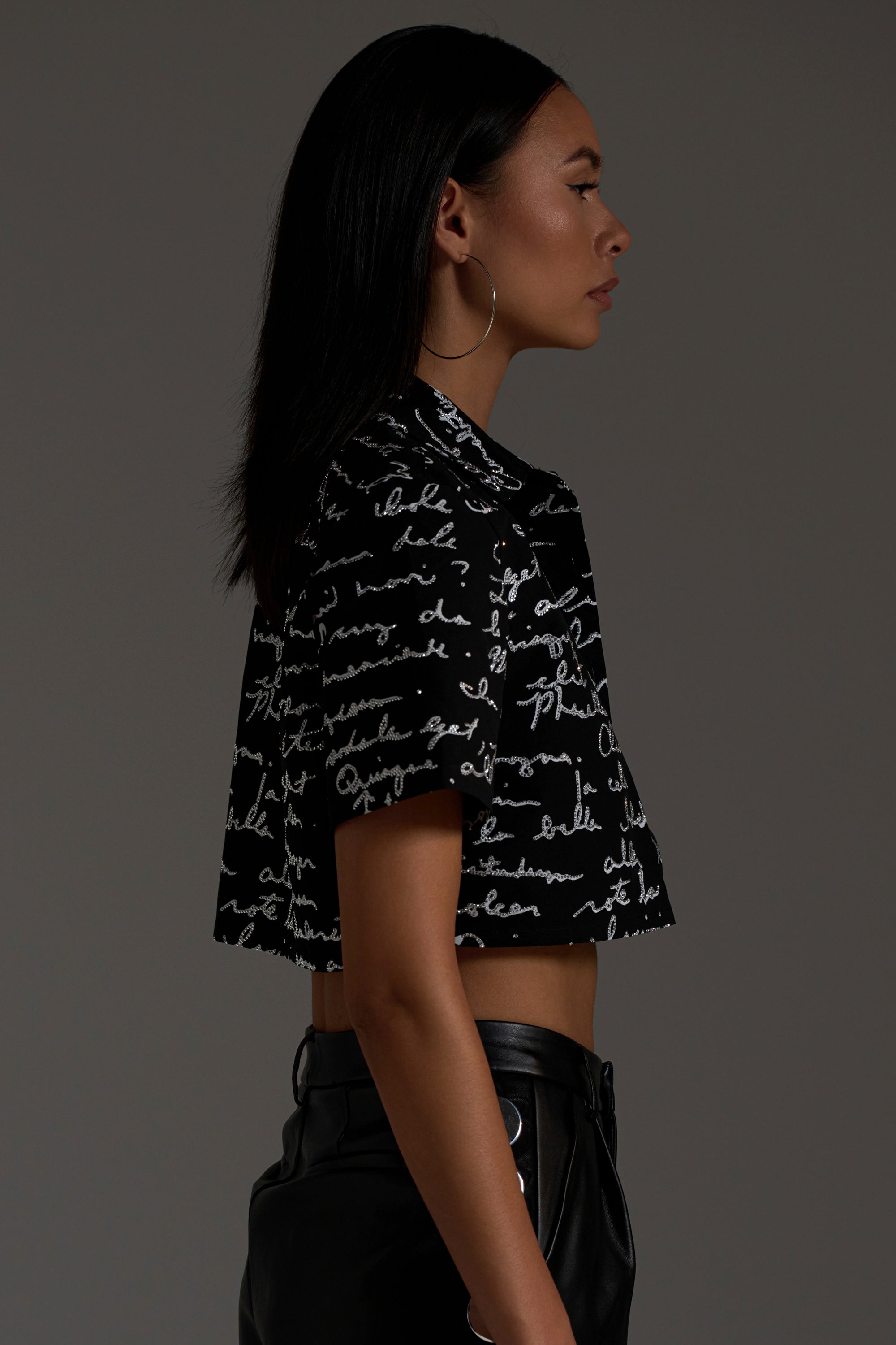 A woman with straight dark hair and hoop earrings wears the CHANGES BUTTON DOWN—a black shirt with white cursive script—and black pants. She is shown in profile against a plain dark background with a neutral expression.