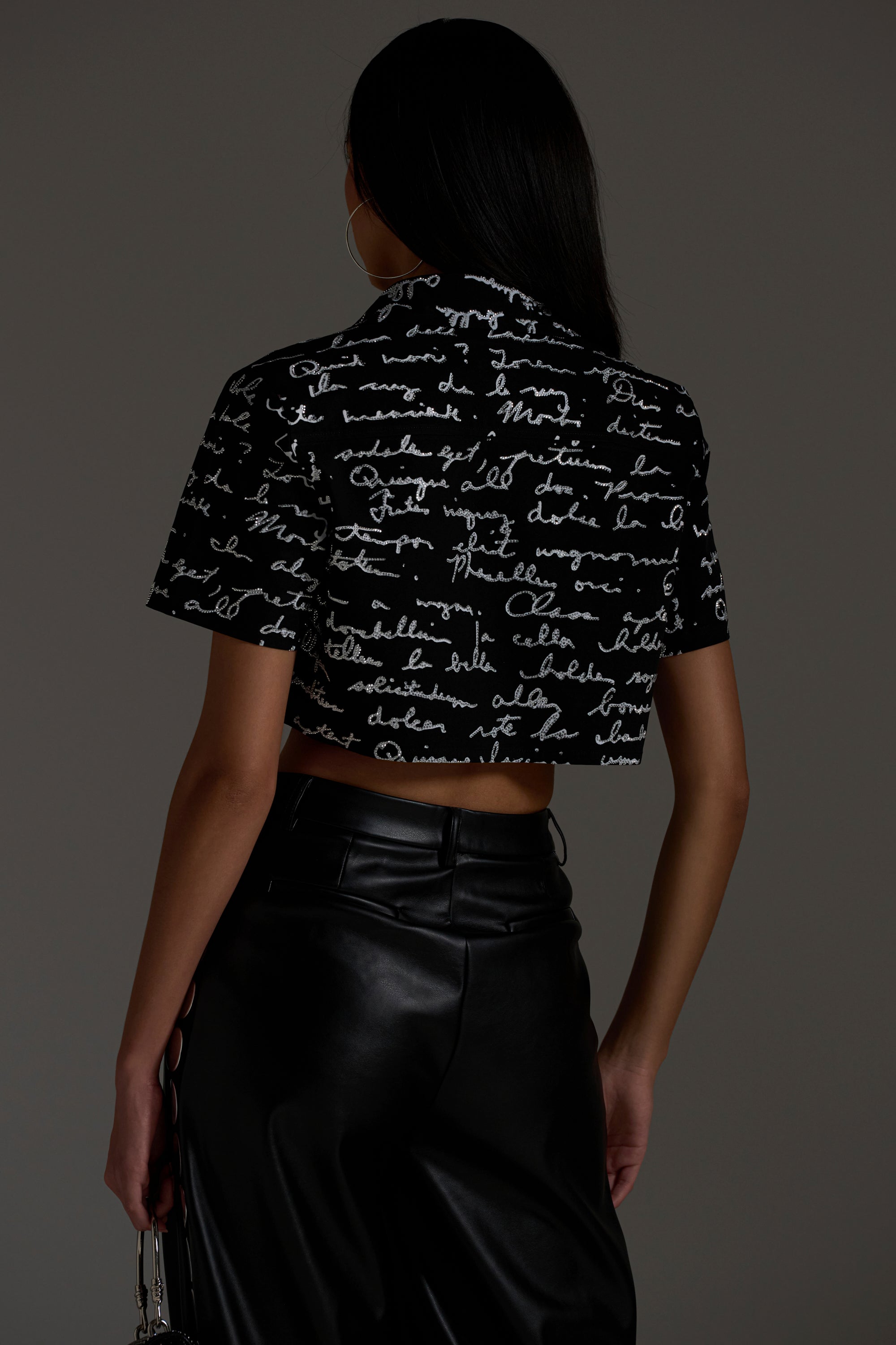 A woman stands with her back to the camera, wearing the CHANGES BUTTON DOWN in black with white cursive writing, paired with high-waisted black leather pants. The boxy silhouette and dark background highlight her striking outline.