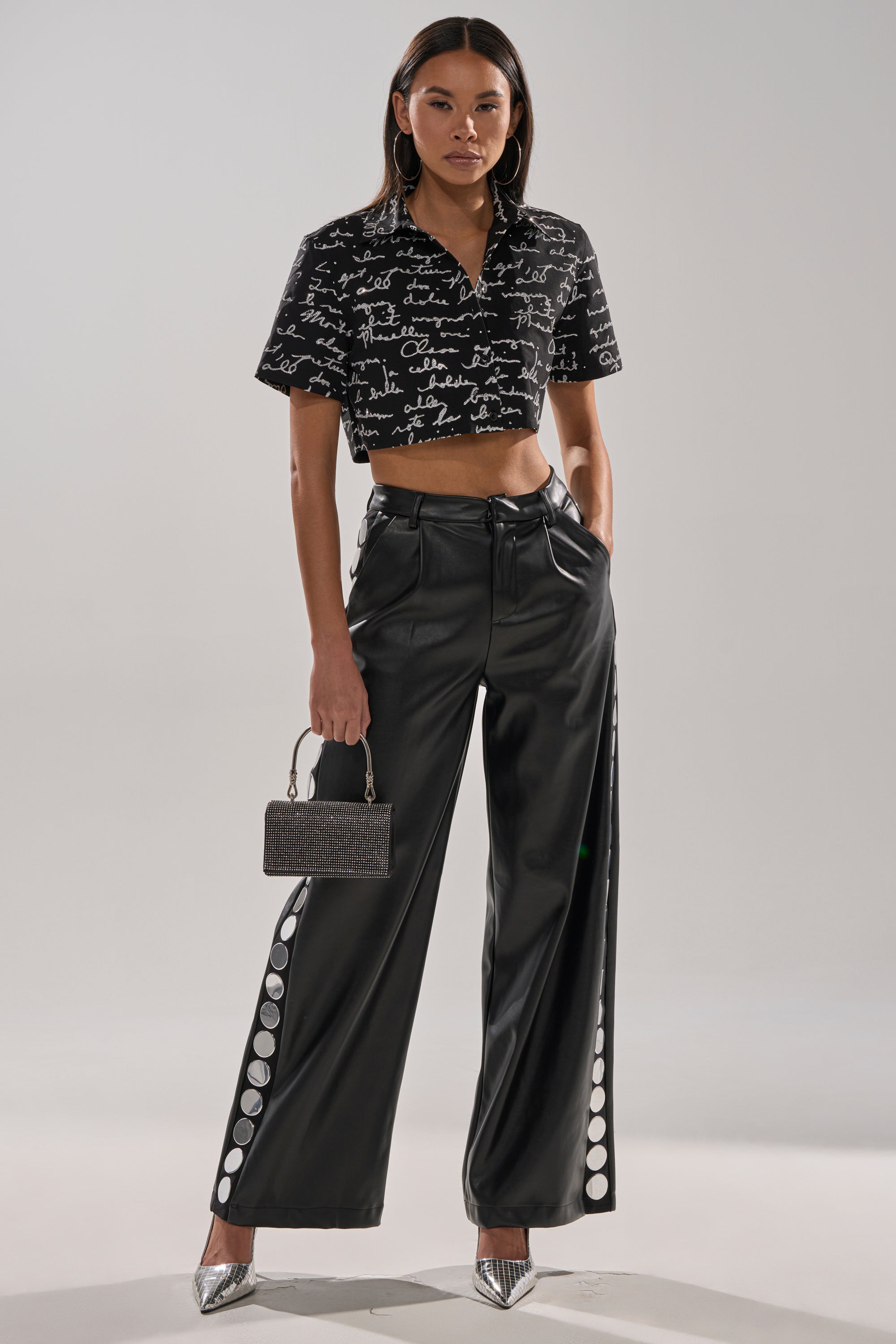 A woman wears the CHANGES BUTTON DOWN, a black script print short-sleeve crop top, paired with high-waisted black wide-leg pants with large side buttons, silver heels, a textured handbag, and has straight dark hair.