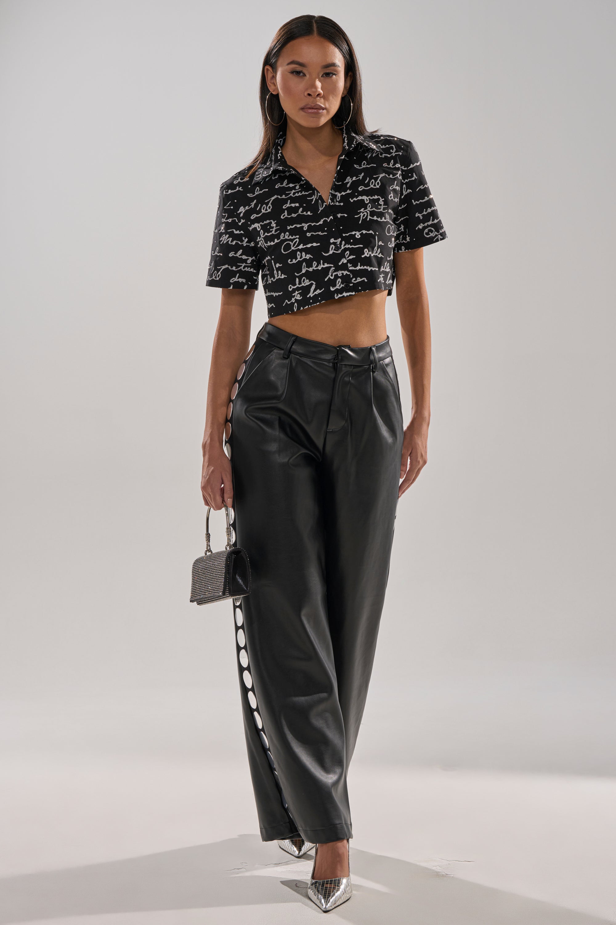 A woman poses in the CHANGES BUTTON DOWN, high-waisted black leather pants with side buttons, metallic pointed-toe heels, and a small silver handbag.