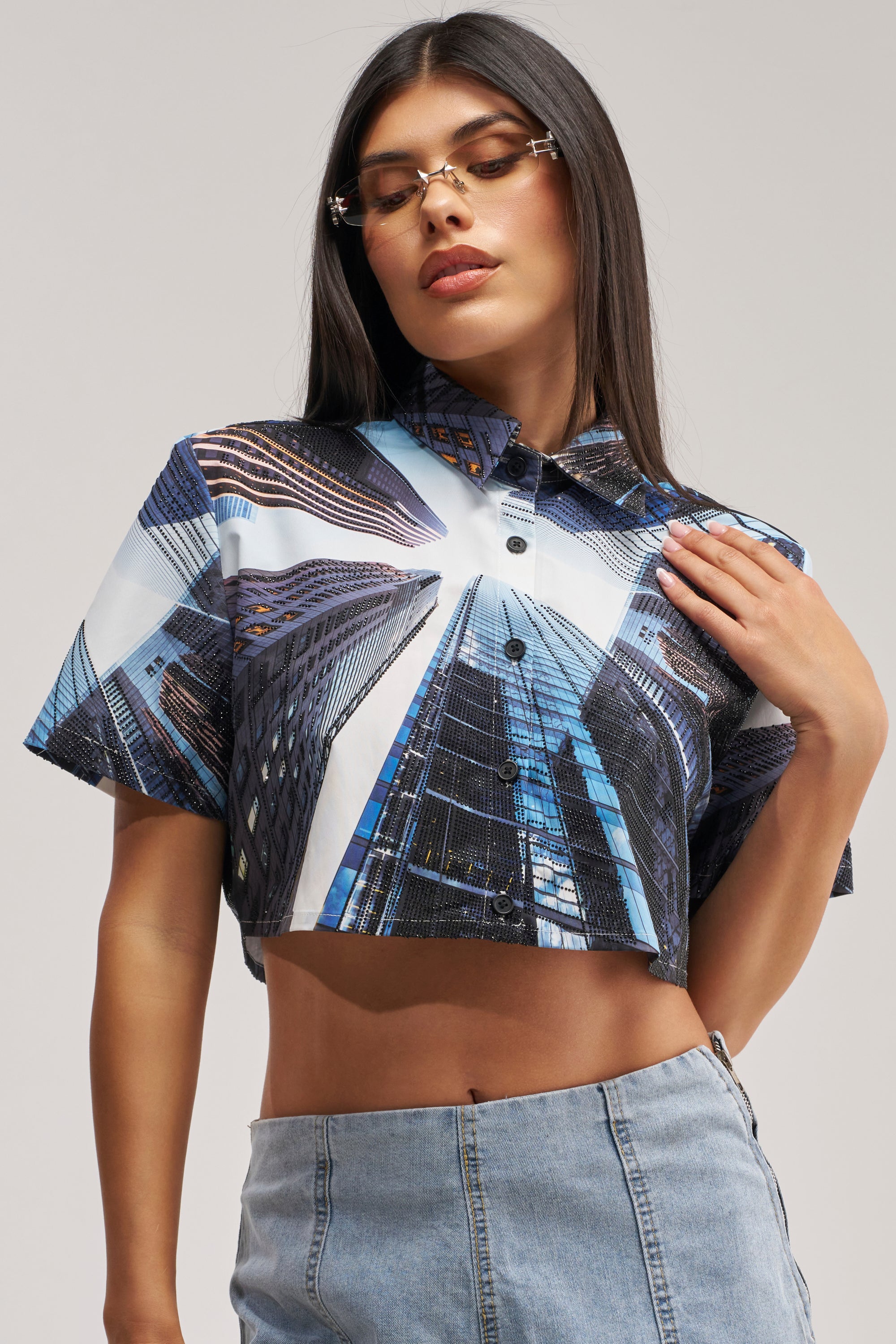 A woman poses against a plain background, wearing the CITY DREAMS POPLIN BLOUSE—a cropped button-up shirt adorned with vivid skyscraper cityscape prints—and pairs it with a light blue denim skirt. She places one hand on her chest while the other rests by her side.