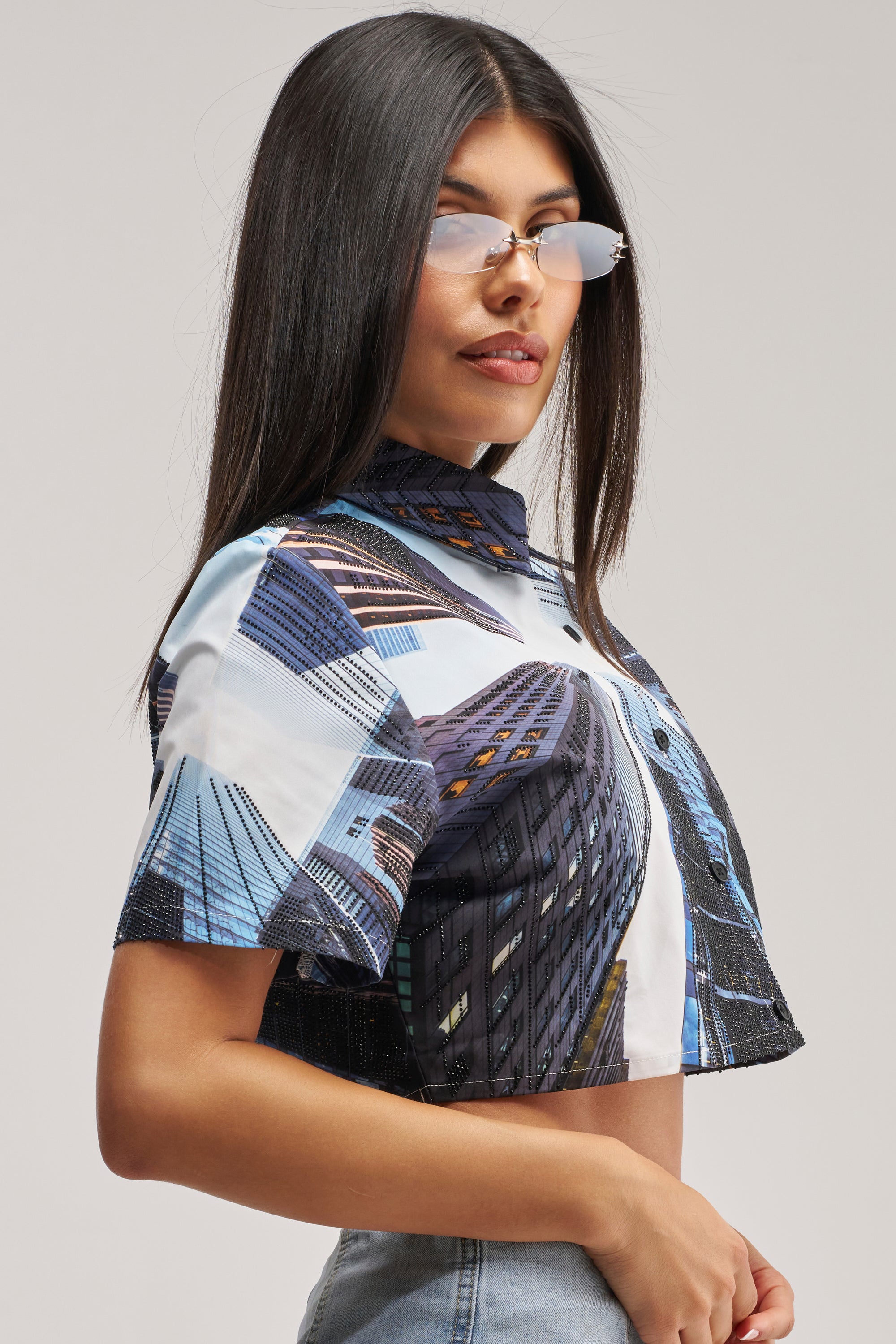 A woman with long dark hair wears reflective sunglasses and the CITY DREAMS POPLIN BLOUSE—a cropped, short-sleeve button-up featuring a bold cityscape print—paired with light denim jeans as she stands against a plain background.