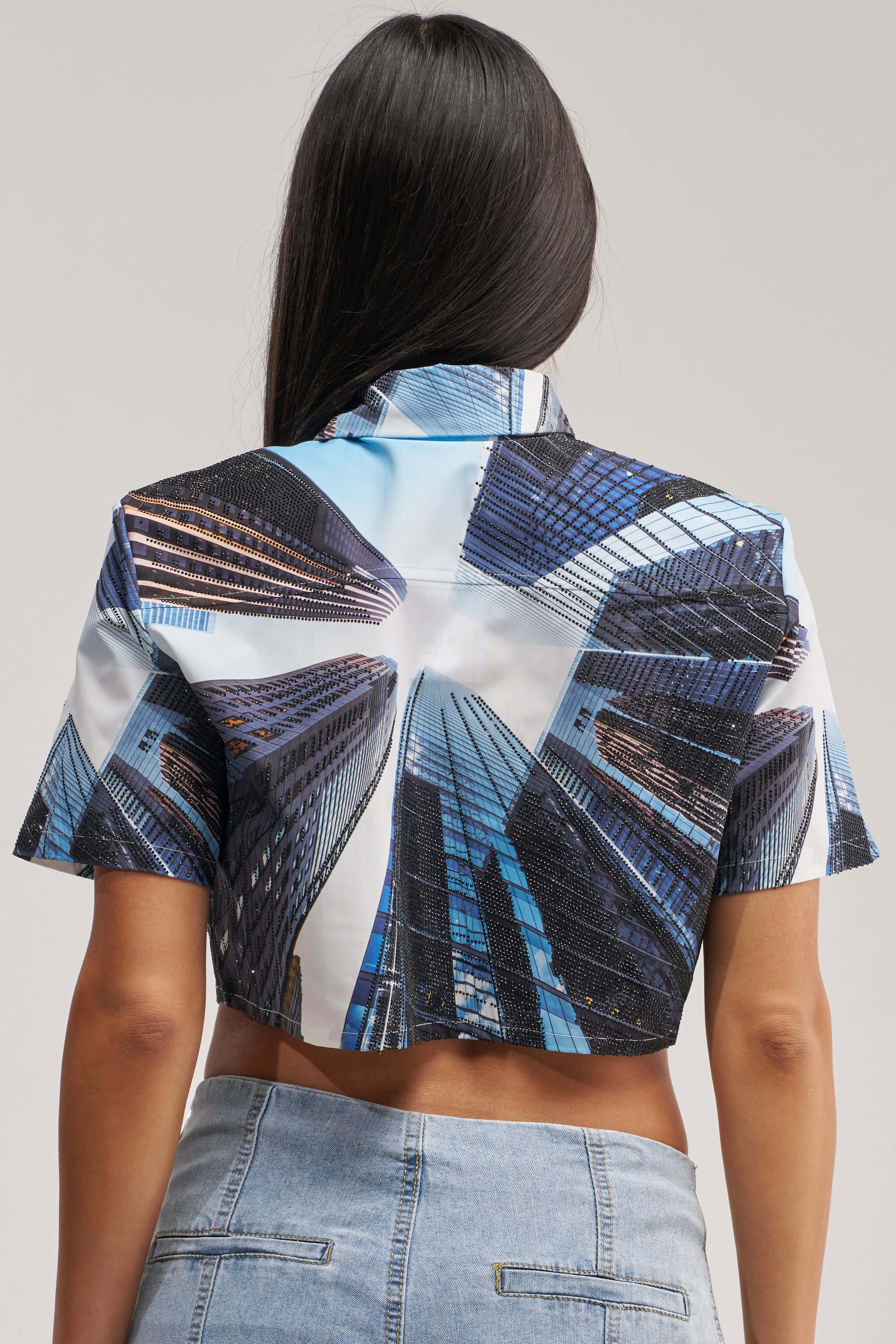 A person with long dark hair is seen from the back, wearing the CITY DREAMS POPLIN BLOUSE—a cropped graphic blouse adorned with a striking cityscape print showing tall buildings beneath a blue sky. They pair it with light denim jeans.