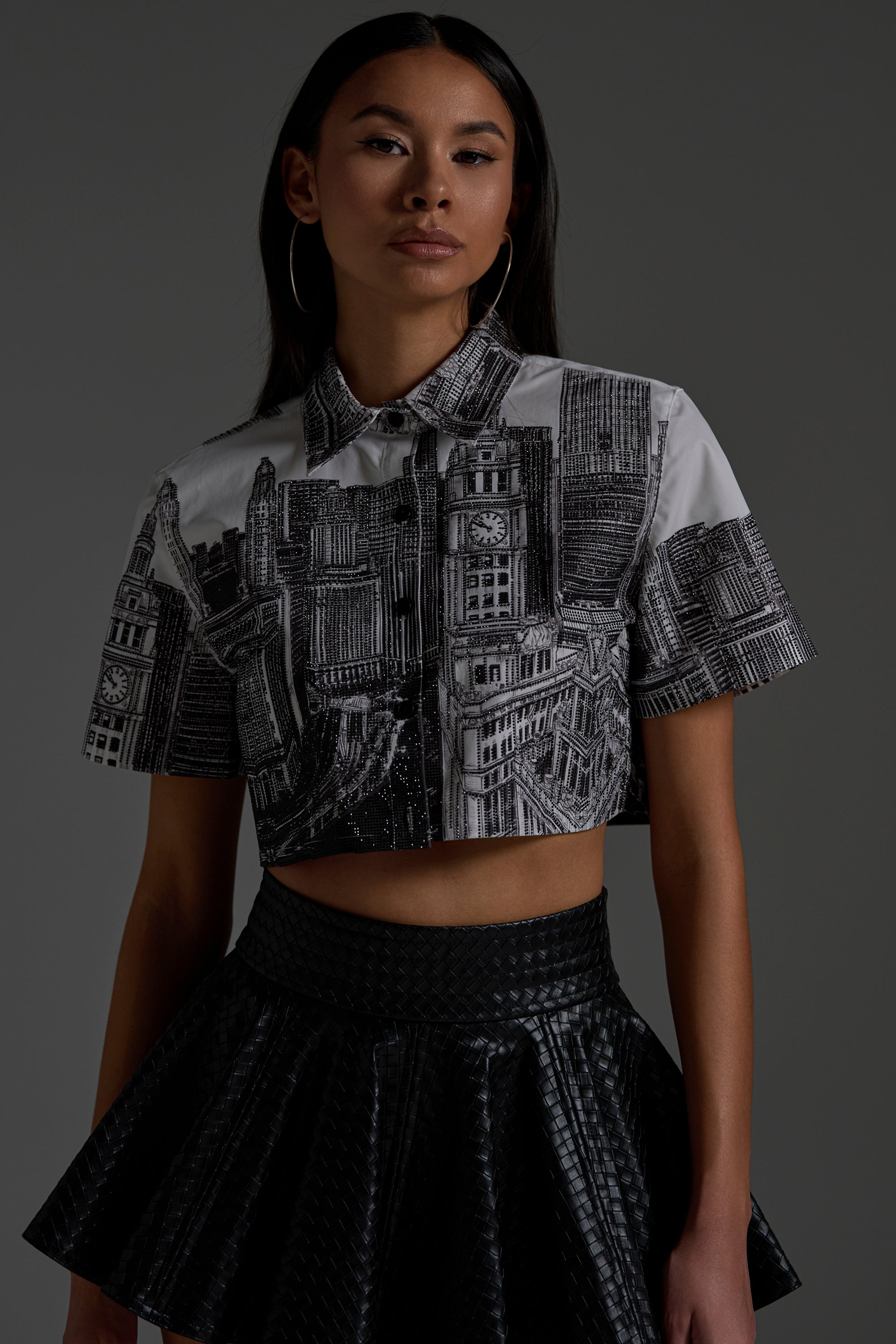 A woman stands against a dark background wearing large hoop earrings, a textured black skirt, and the ALL OVER THE CITY BUTTON DOWN styled as a cropped shirt with a cityscape print.