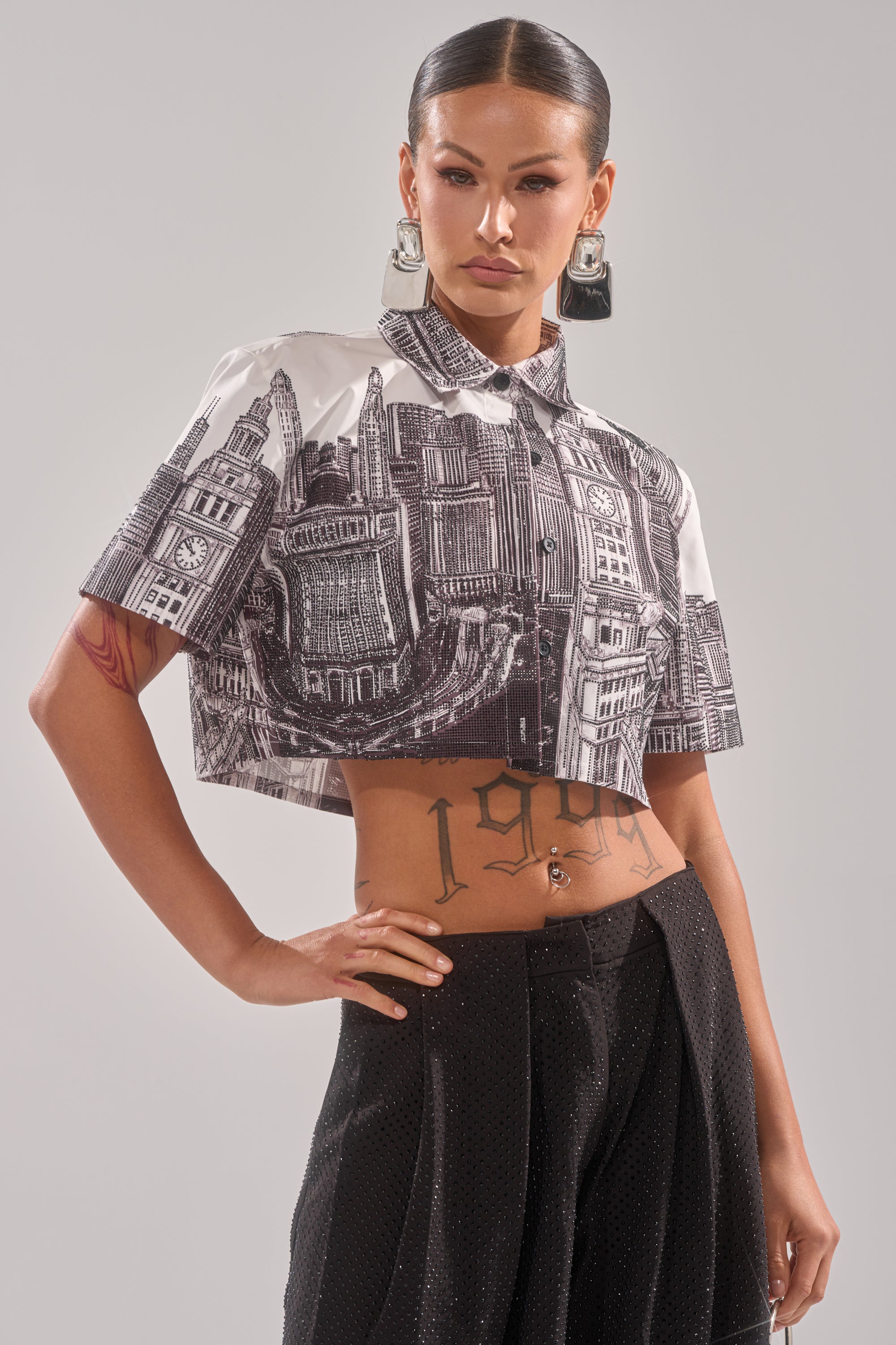 A woman with slicked-back hair wears the ALL OVER THE CITY BUTTON DOWN—a rhinestone-adorned black and white cityscape print shirt—paired with dark high-waisted pants, bold statement earrings, and displays a "1994" tattoo on her lower abdomen. 0