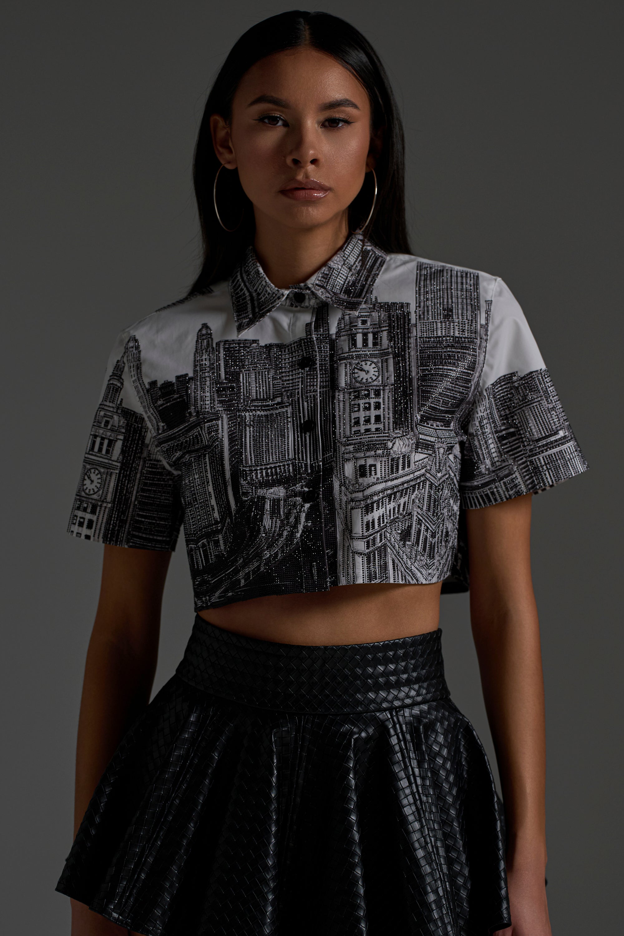 A woman with long dark hair and hoop earrings wears the ALL OVER THE CITY BUTTON DOWN and a black textured flared skirt, standing against a plain gray background.