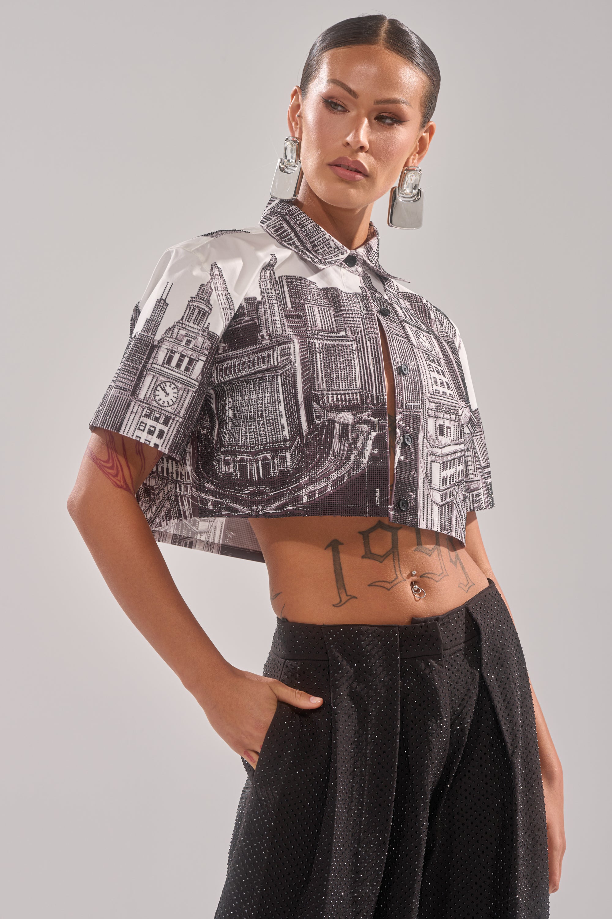A woman poses confidently in the ALL OVER THE CITY BUTTON DOWN, a cropped shirt with a black and white cityscape print, styled with black wide-leg pants, large silver earrings, and showcasing tattoos on her torso and arm. 1