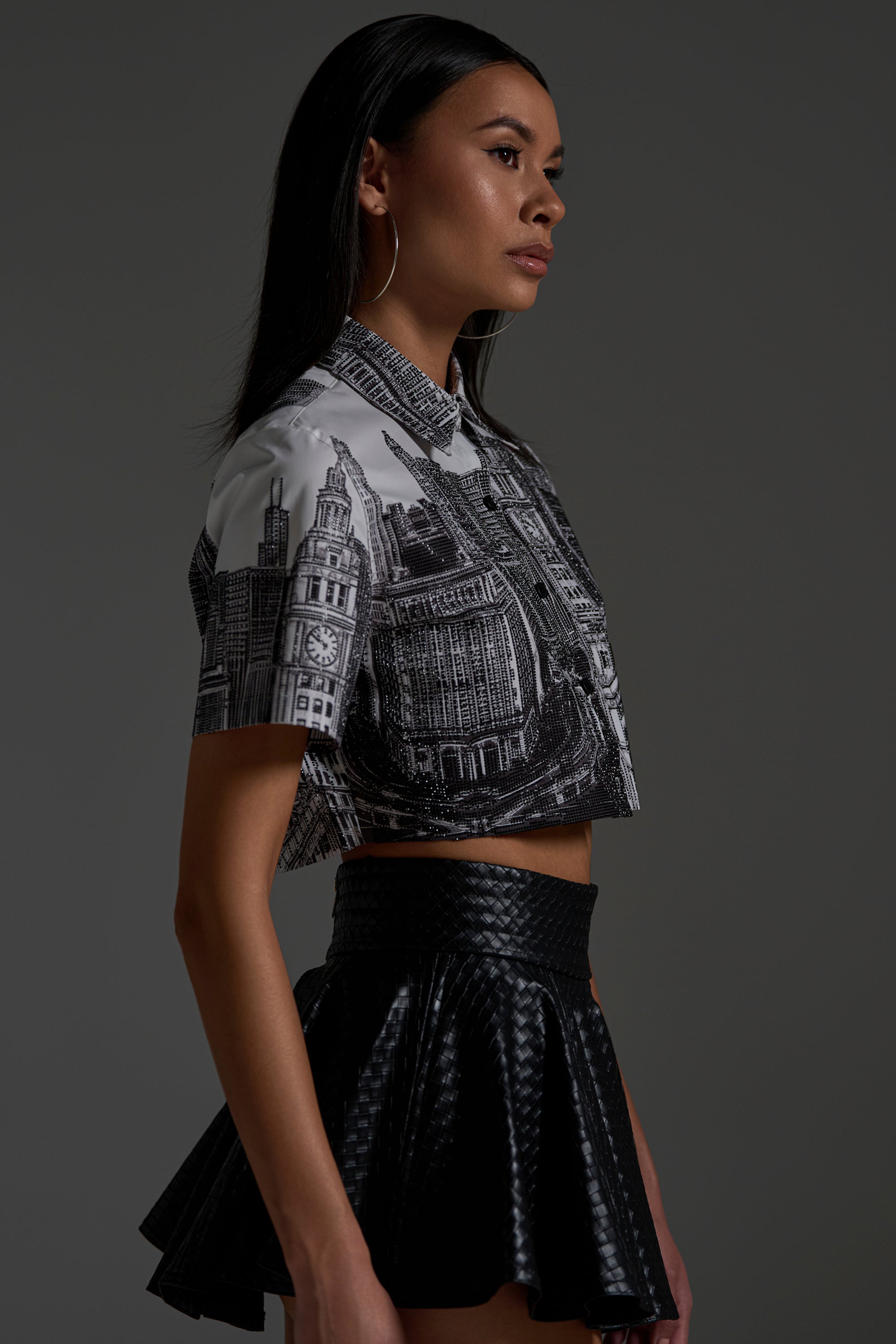 A woman in profile, with long straight hair and large hoop earrings, wears the ALL OVER THE CITY BUTTON DOWN styled as a black-and-white crop top and pairs it with a black, textured, high-waisted mini skirt against a gray background.