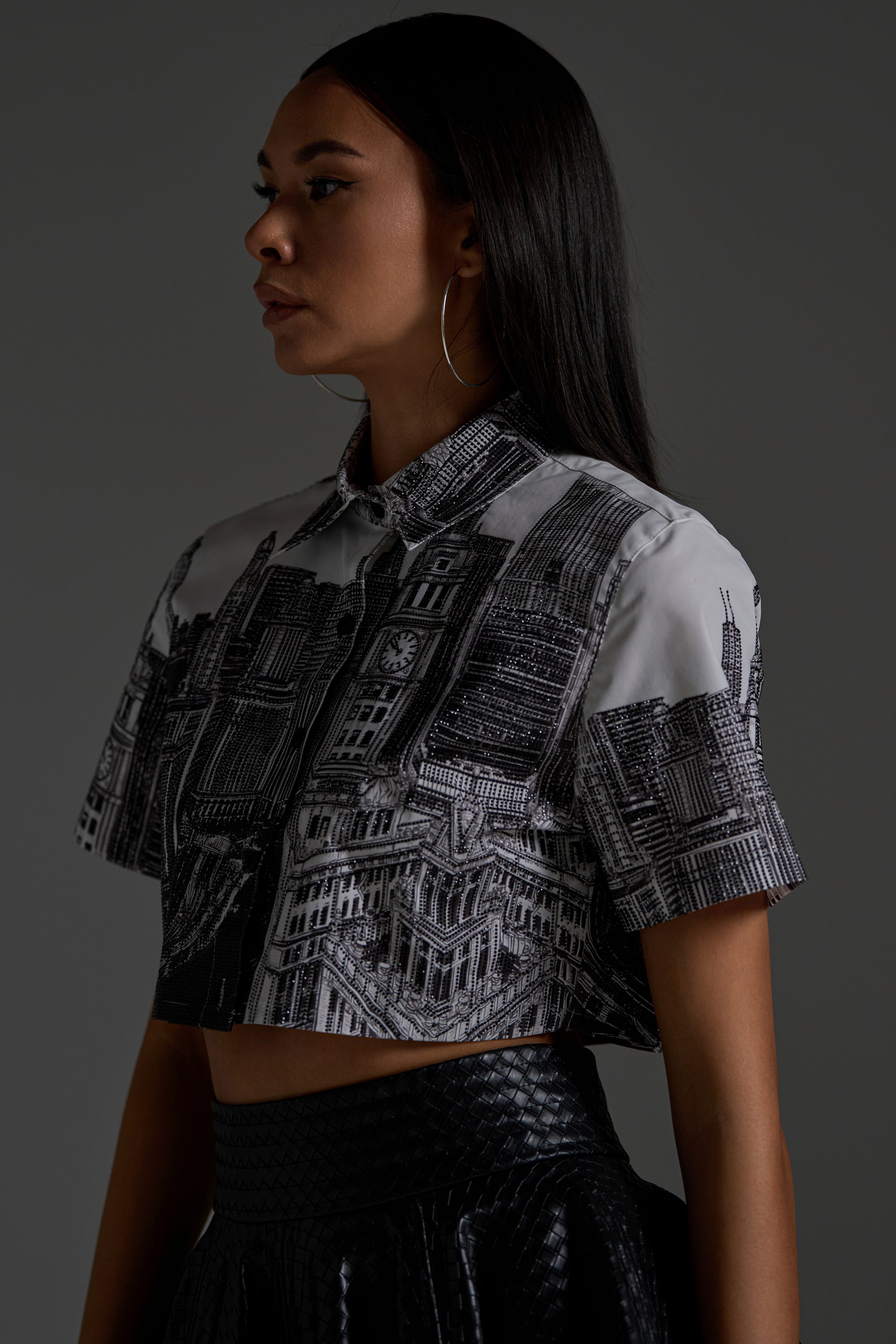 A woman with long dark hair and hoop earrings wears the ALL OVER THE CITY BUTTON DOWN, a cropped shirt with a detailed cityscape print, paired with a textured black skirt against a plain dark background.