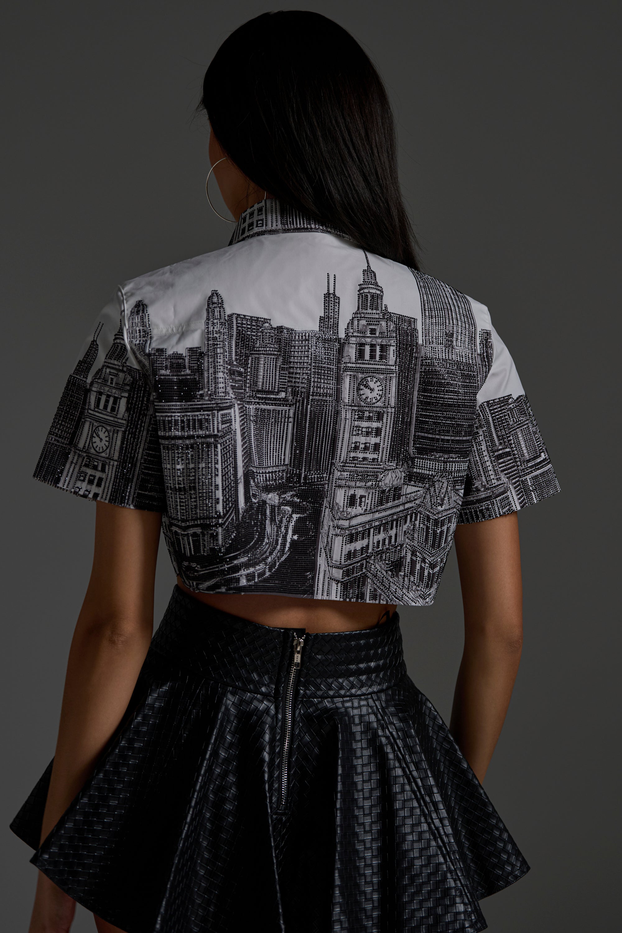 A person with long dark hair faces away, wearing the ALL OVER THE CITY BUTTON DOWN shirt styled cropped and a high-waisted textured black skirt with a back zipper, set against a plain gray background.