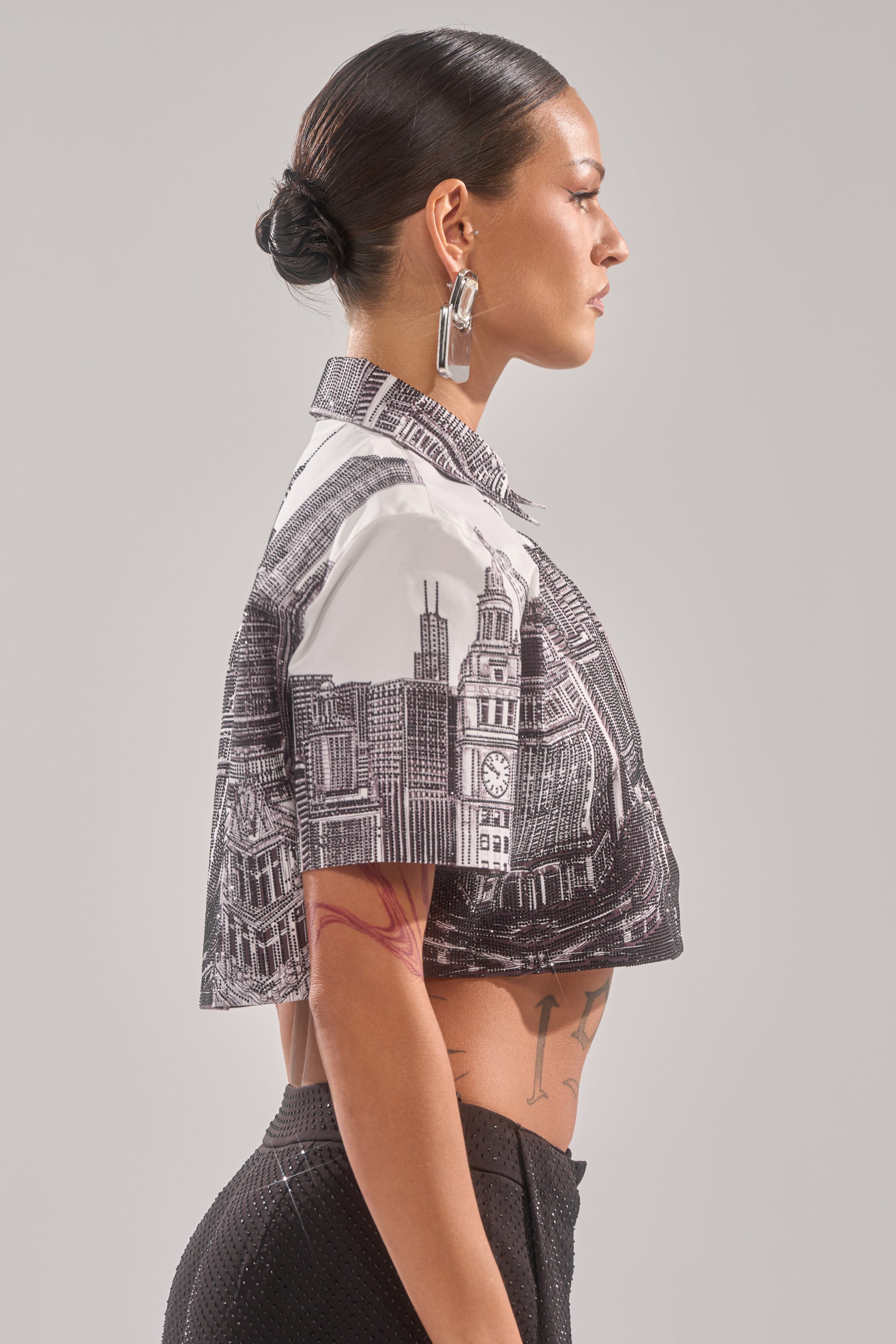 A woman with dark hair in a low bun, wearing geometric earrings and the ALL OVER THE CITY BUTTON DOWN, stands in profile against a light gray background. 4
