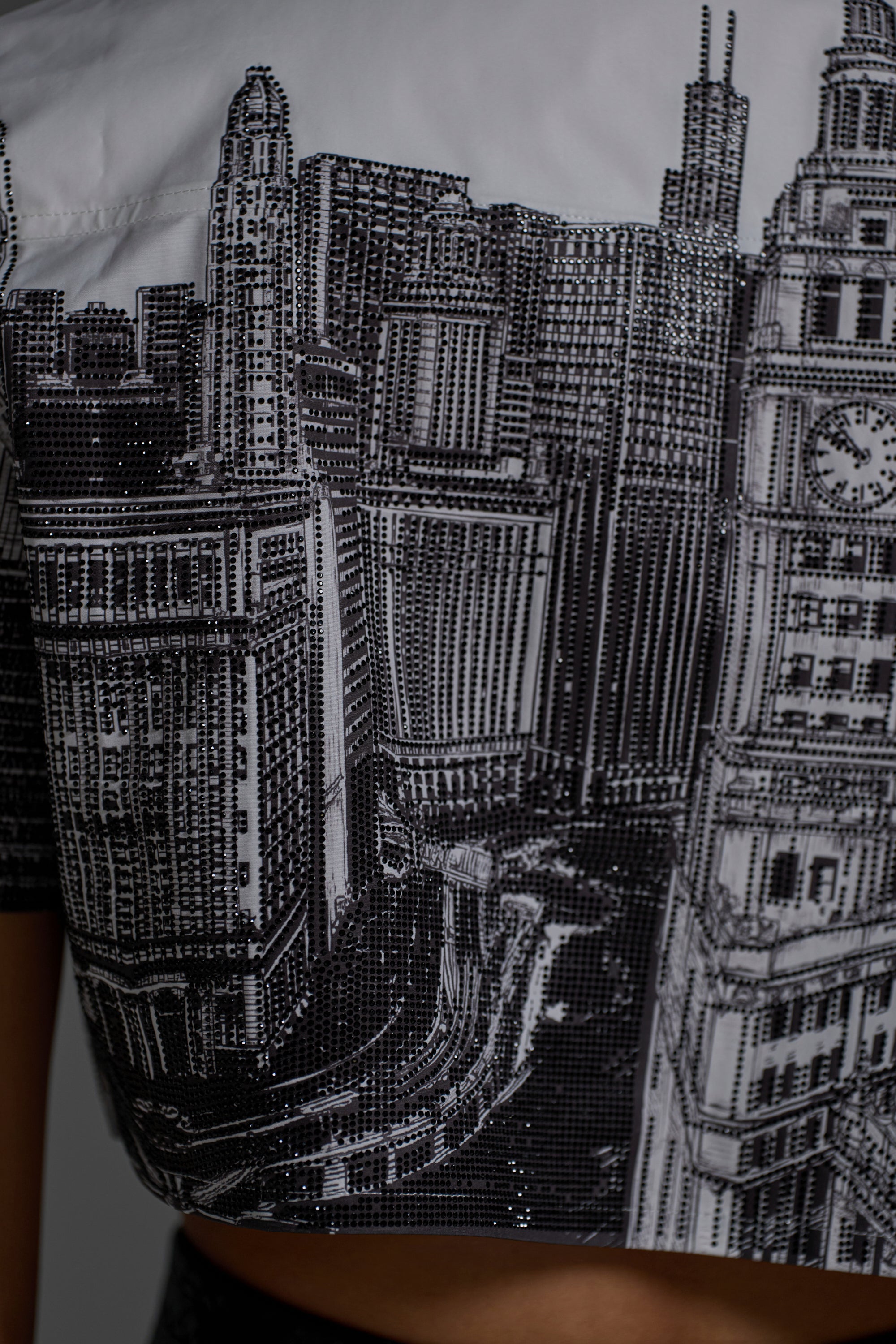 The ALL OVER THE CITY BUTTON DOWN is a trendy cropped shirt featuring a detailed black and white cityscape print with tall buildings and a clock tower.