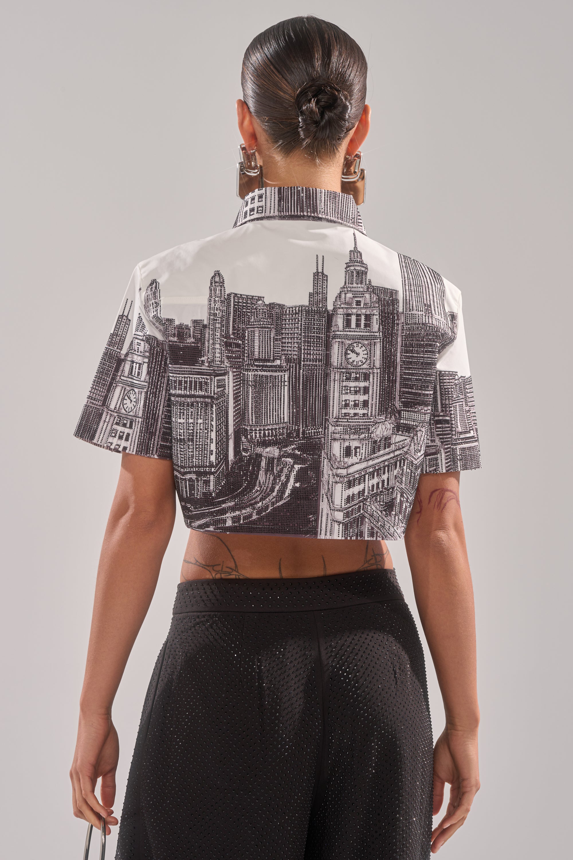 A woman with her hair in a sleek bun faces away from the camera, wearing the ALL OVER THE CITY BUTTON DOWN shirt, paired with black textured pants and holding a small object in her right hand. 5