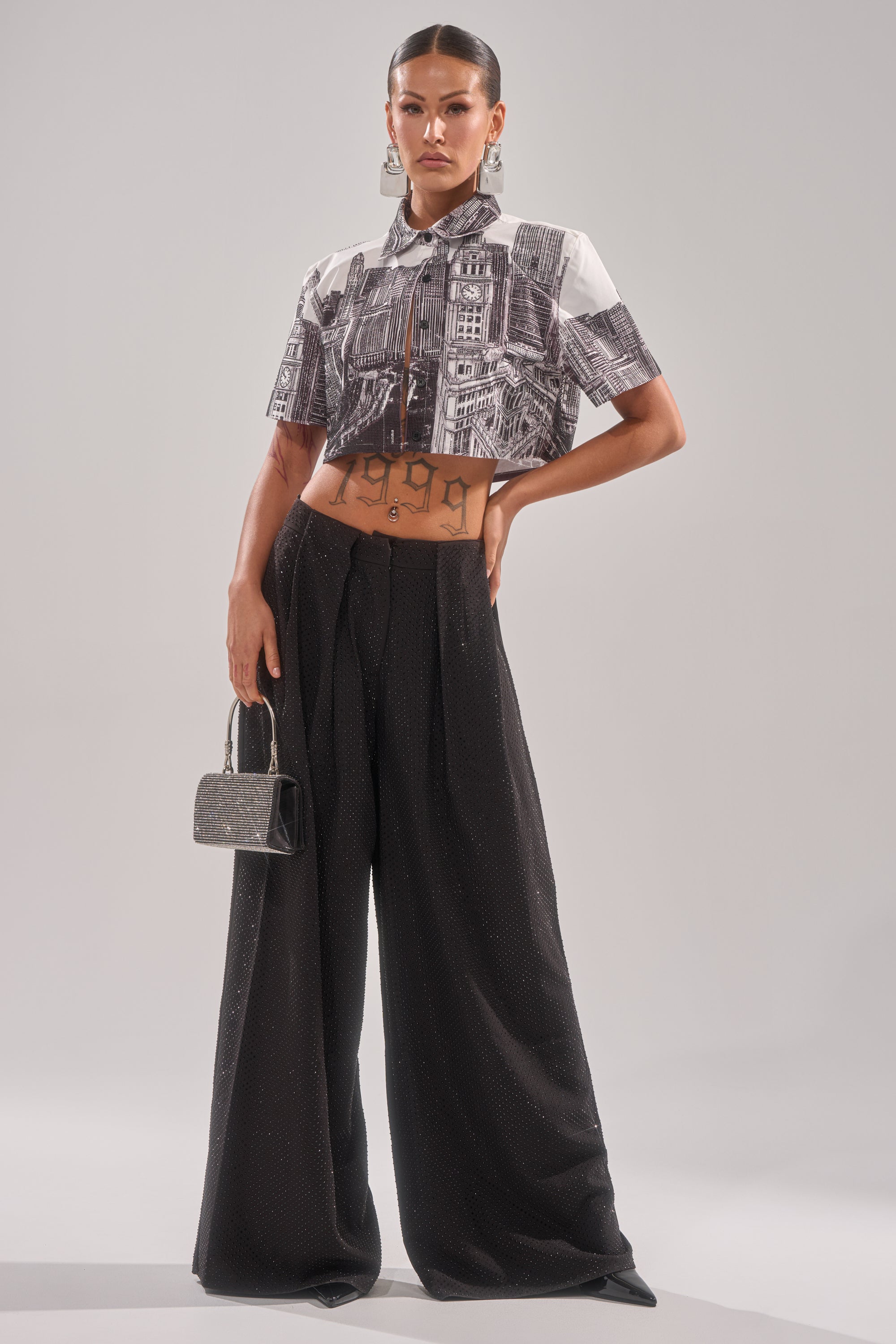 A person stands confidently in the ALL OVER THE CITY BUTTON DOWN, a cropped cityscape print shirt, paired with high-waisted wide-leg black pants. They accessorize with a small metallic handbag and large hoop earrings. 6
