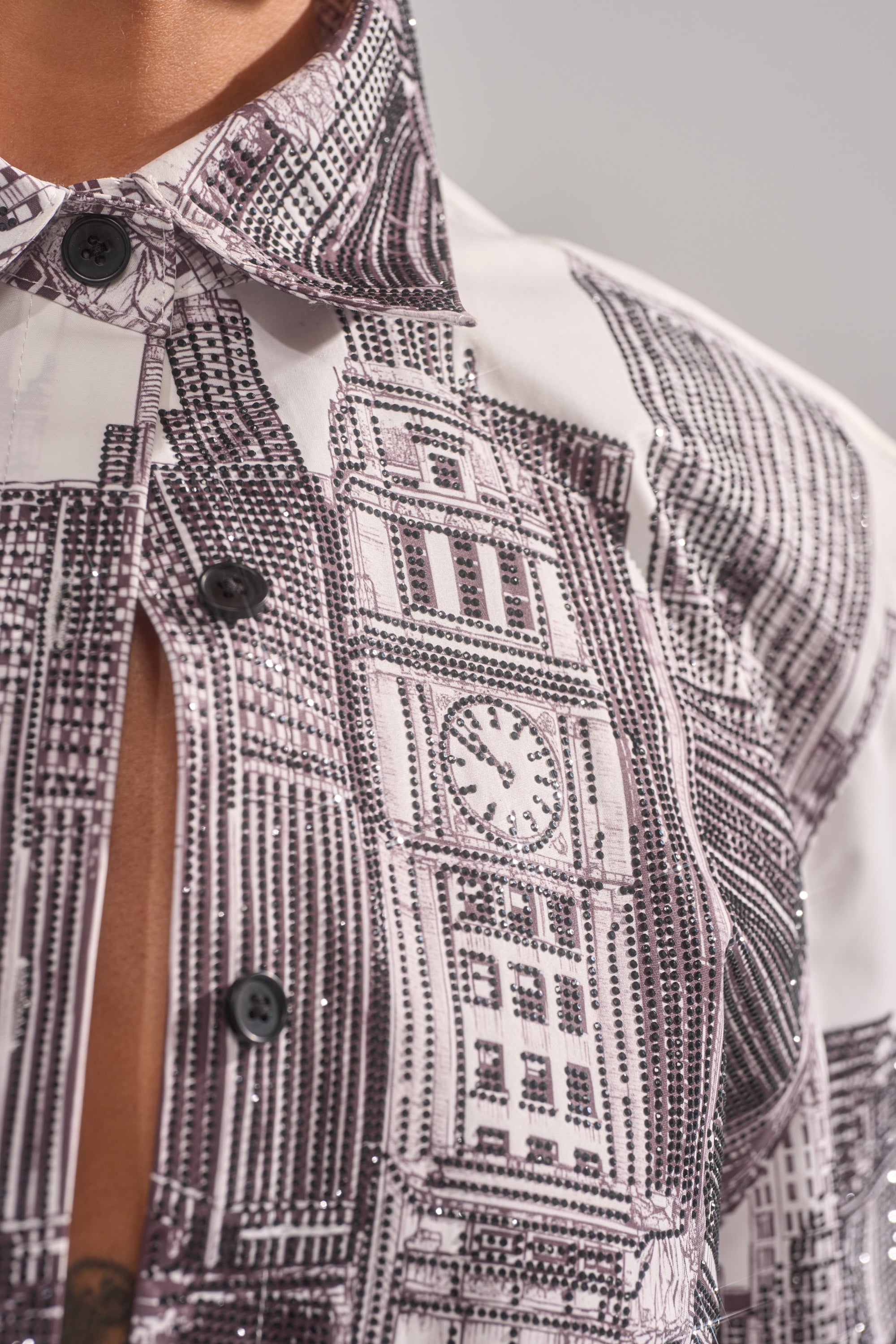 Close-up of the ALL OVER THE CITY BUTTON DOWN, a white shirt with black buttons and a cityscape print featuring architectural details and a clock tower. Shirt is slightly open at the chest. 8
