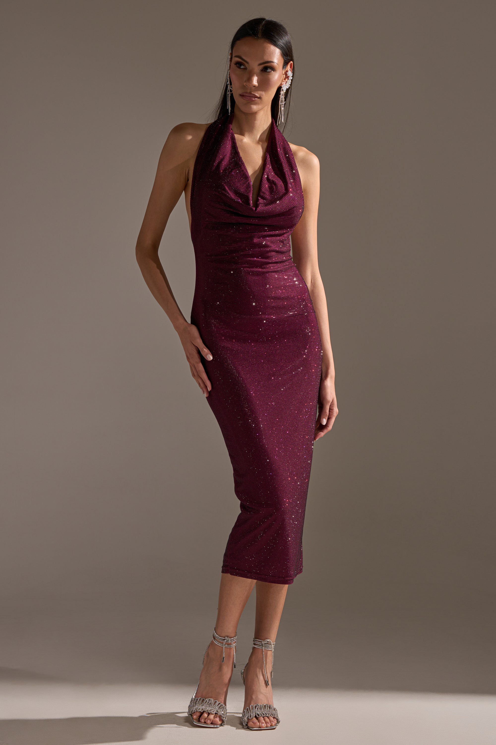 A woman poses in the POPPIN' BOTTLES RHINESTONE MAXI DRESS IN PURPLE, a sleeveless sparkly halter style. She has long dark hair and accessorizes with silver dangling earrings and silver high-heeled sandals against a plain background.