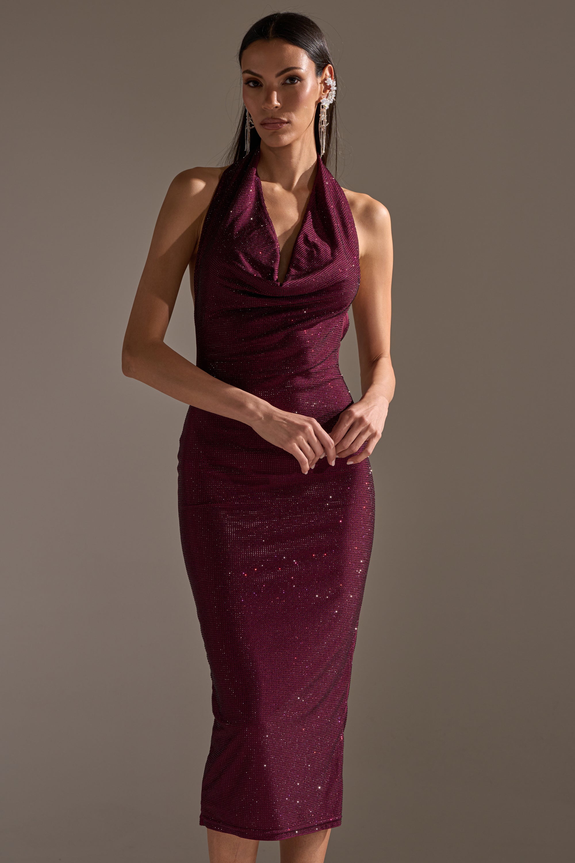 A woman stands against a plain background in the POPPIN' BOTTLES RHINESTONE MAXI DRESS IN PURPLE—a sleeveless, sparkly halter maxi dress with a draped neckline. She has long straight hair and wears statement earrings to complete her look.