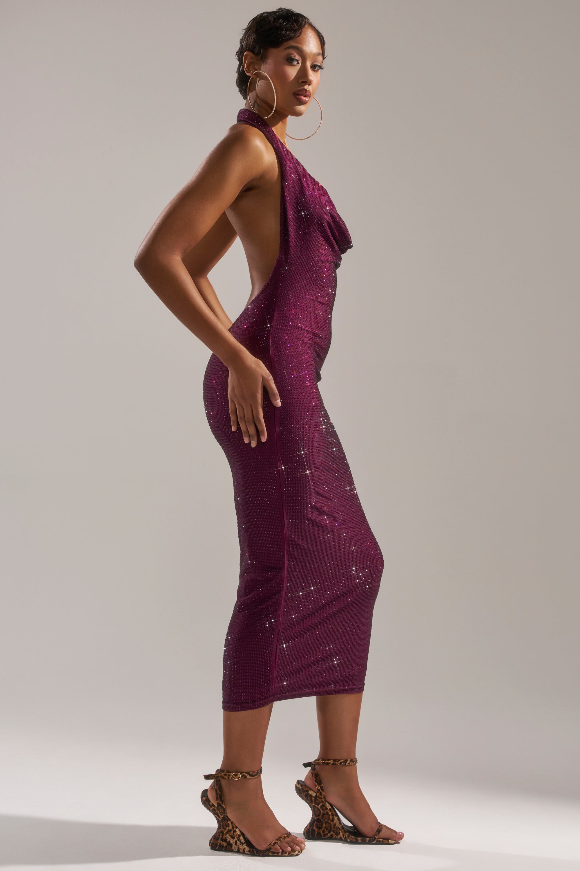 A woman poses in profile wearing the POPPIN' BOTTLES RHINESTONE MAXI DRESS IN PURPLE, a dazzling sleeveless and backless evening gown, paired with leopard-print high-heeled sandals against a light plain background.