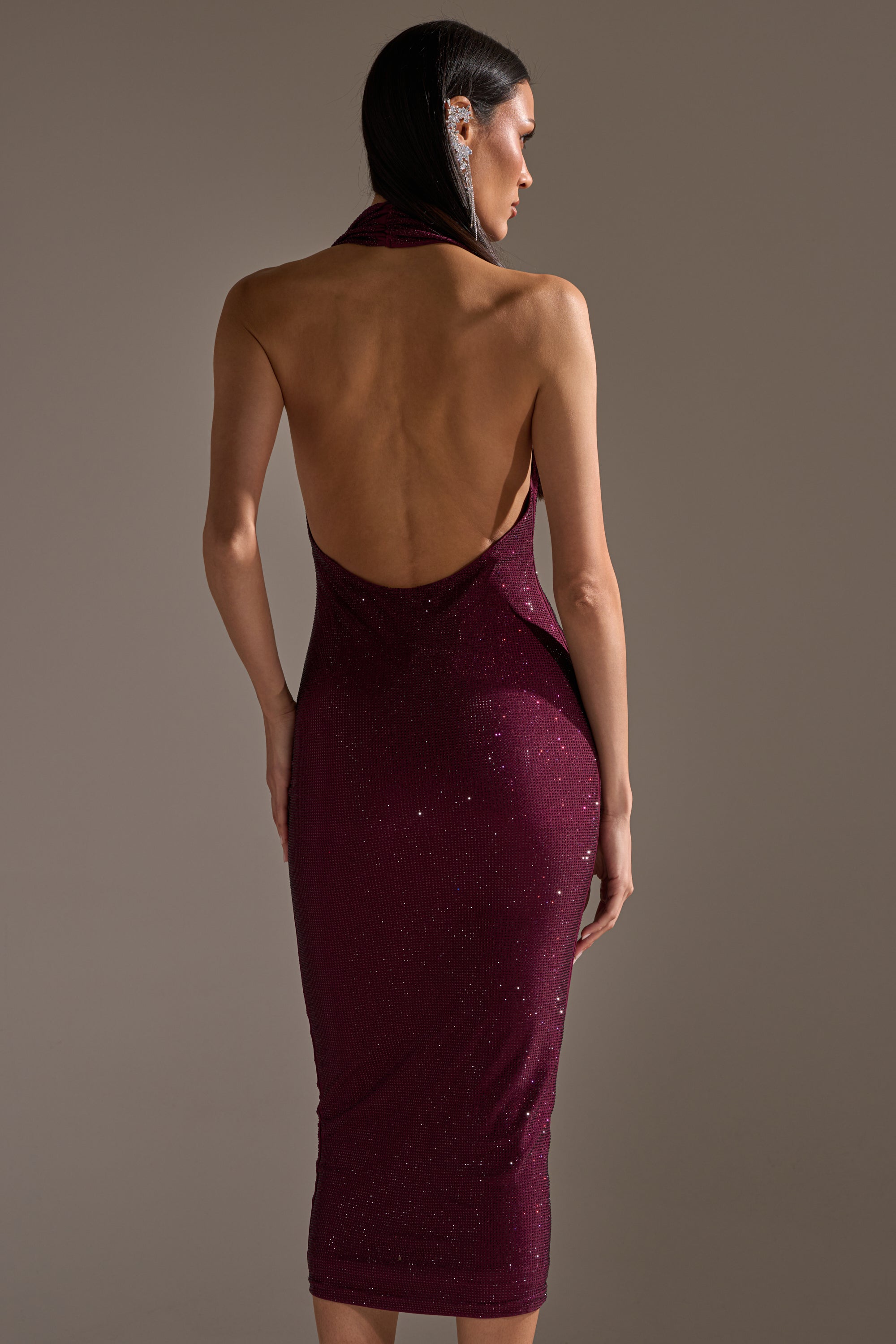 A woman poses with her back to the camera in the POPPIN' BOTTLES RHINESTONE MAXI DRESS IN PURPLE, a sparkling, sleeveless, backless, form-fitting gown. Her straight dark hair drapes over one shoulder against a plain background.