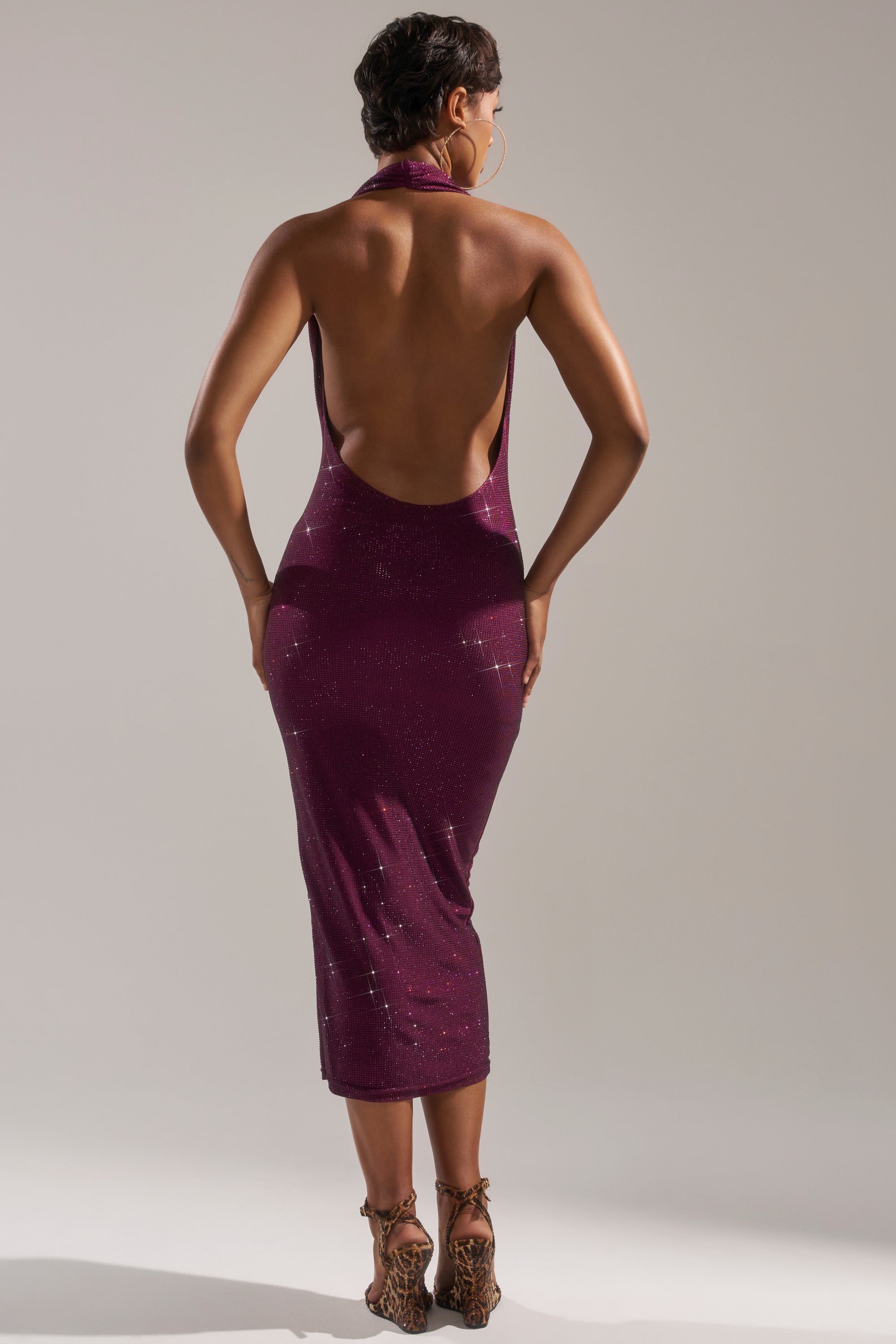 A person stands facing away, wearing the POPPIN' BOTTLES RHINESTONE MAXI DRESS IN PURPLE, a sparkly, backless halter-neck evening gown paired with leopard print high heels. The dress pops against the plain, light-colored background.