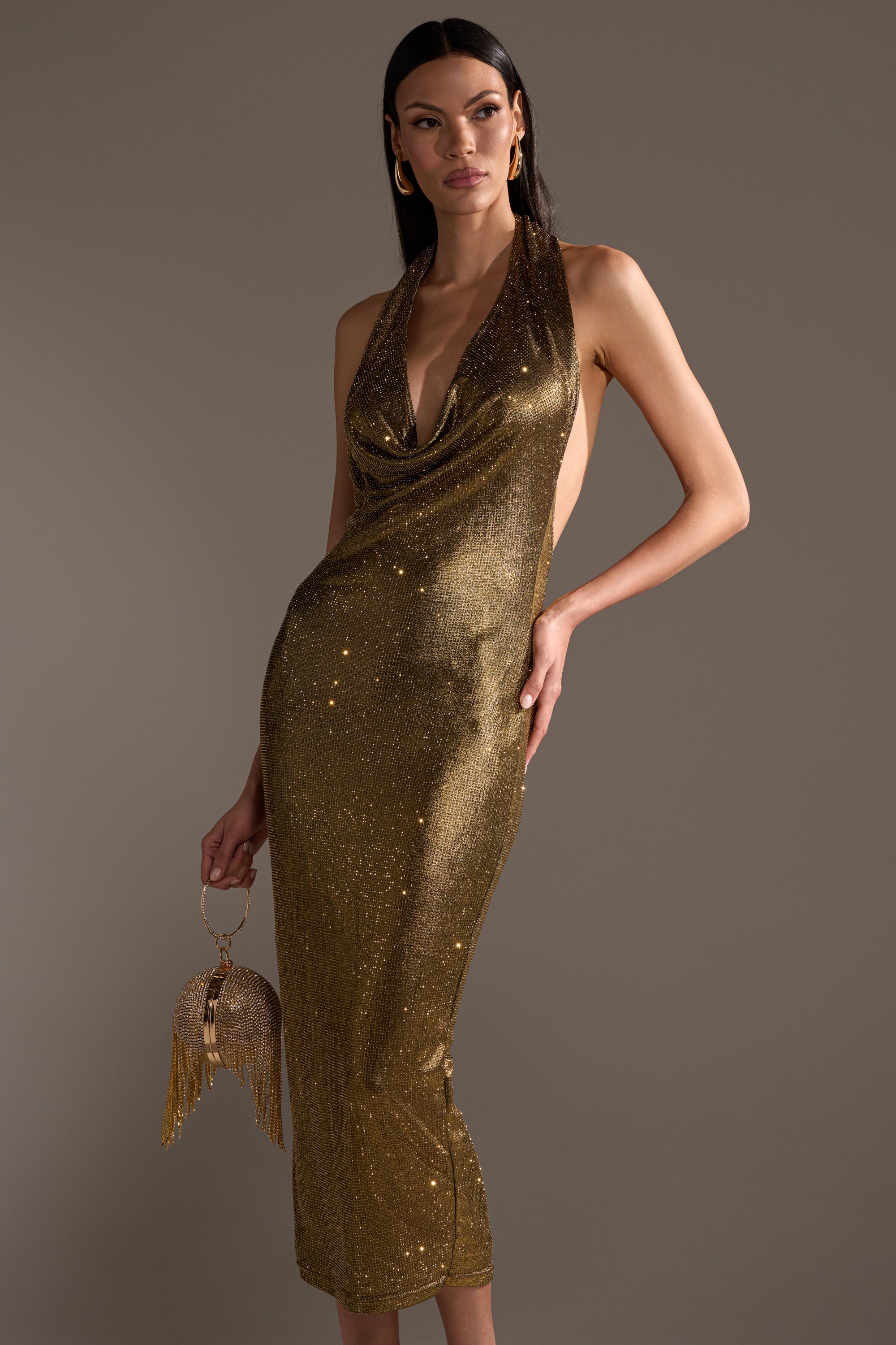 A woman poses in the POPPIN' BOTTLES RHINESTONE MAXI DRESS IN GOLD, featuring a shimmering curve-hugging silhouette, draped neckline, and backless halter style. She accessorizes with a gold fringed purse and hoop earrings against a neutral background.