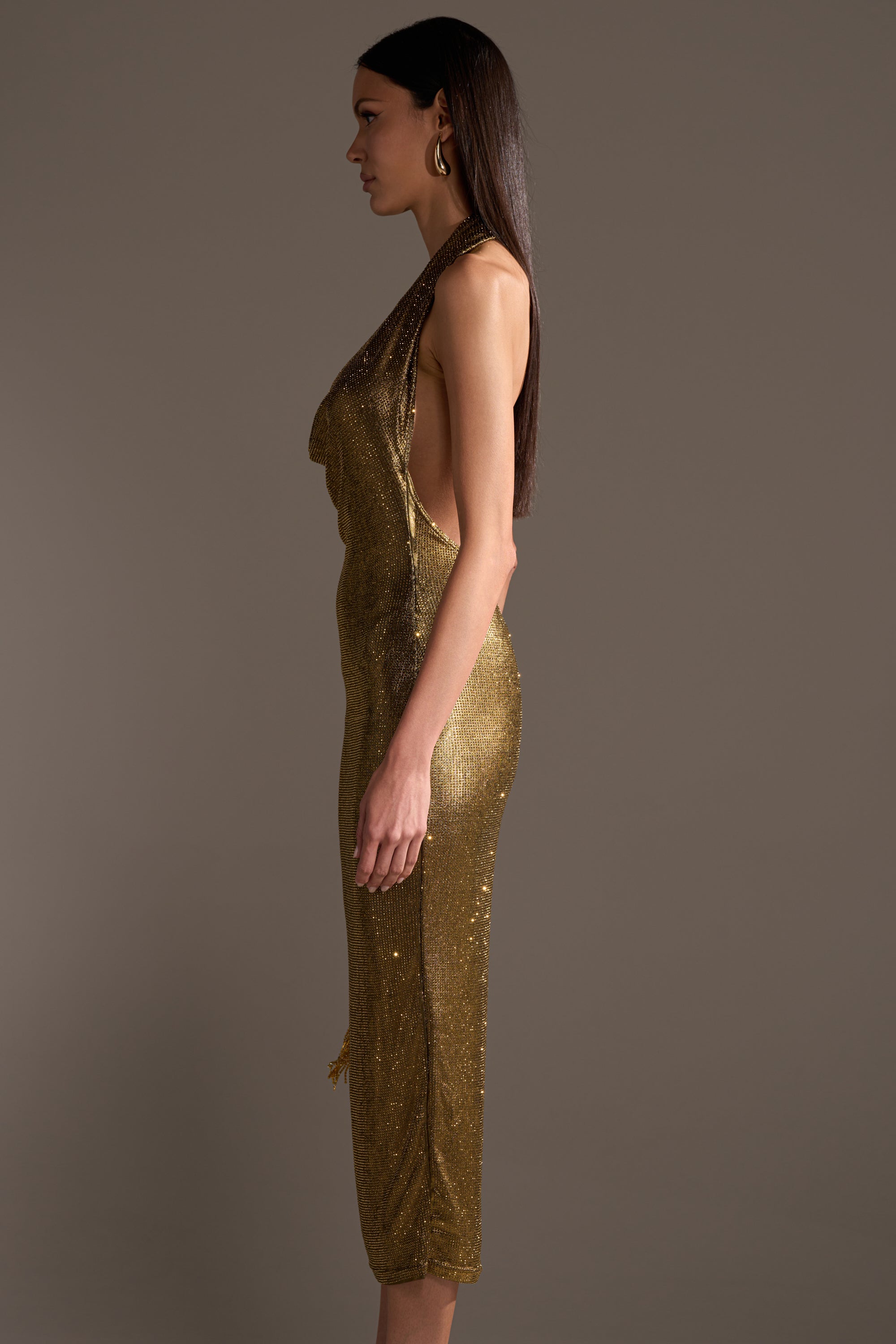 A woman with long straight hair stands in profile against a plain background, wearing the POPPIN' BOTTLES RHINESTONE MAXI DRESS IN GOLD—a sleeveless, open-back, fitted party dress with shimmering gold rhinestones and a striking calf-length silhouette.