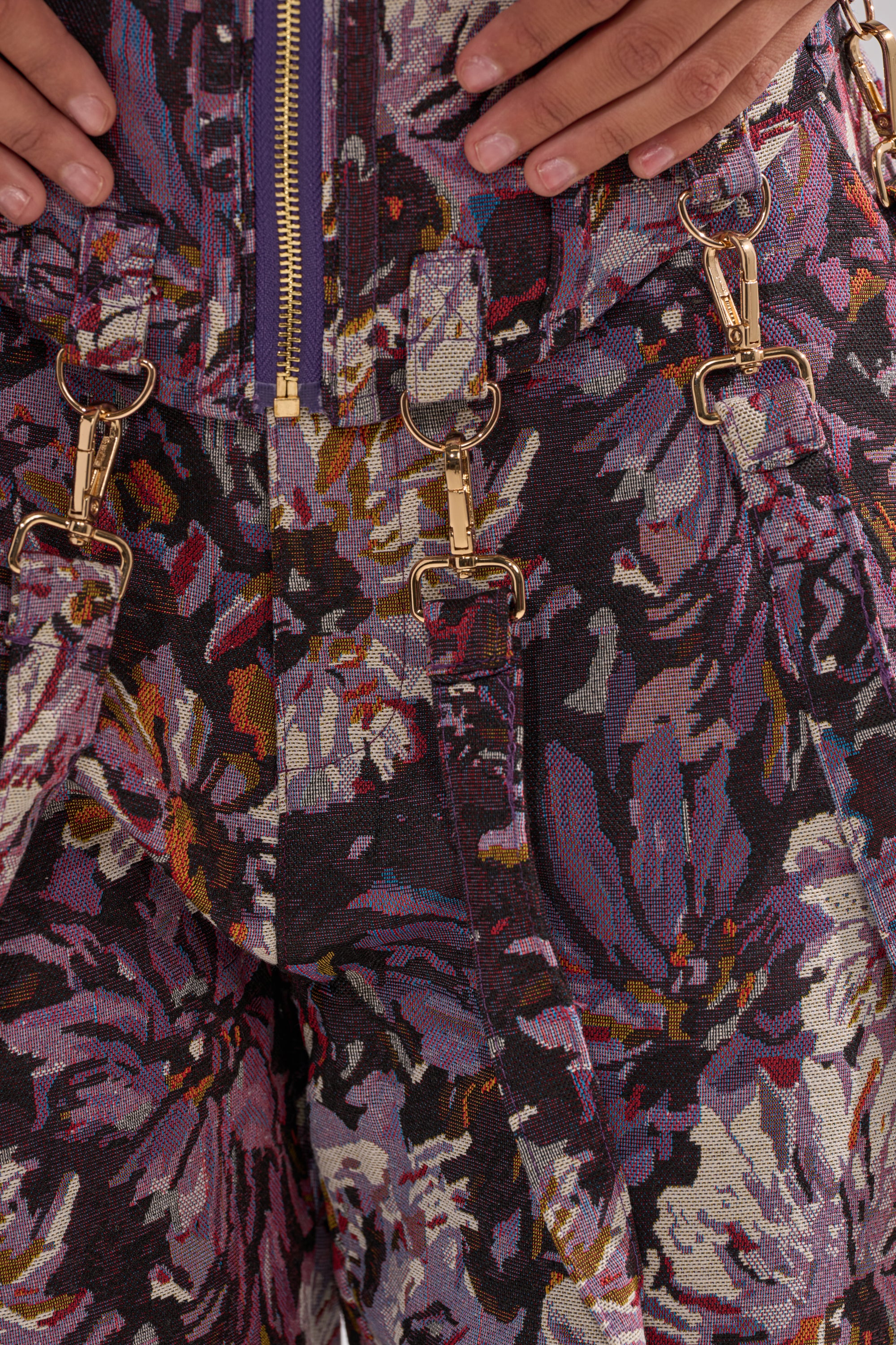 Close-up of the ABOVE THE MIDDLE FLORAL CORSET in purple, pink, and gold jacquard with gold metal clip fasteners and a visible purple zipper. Two hands rest near the top of the corset.