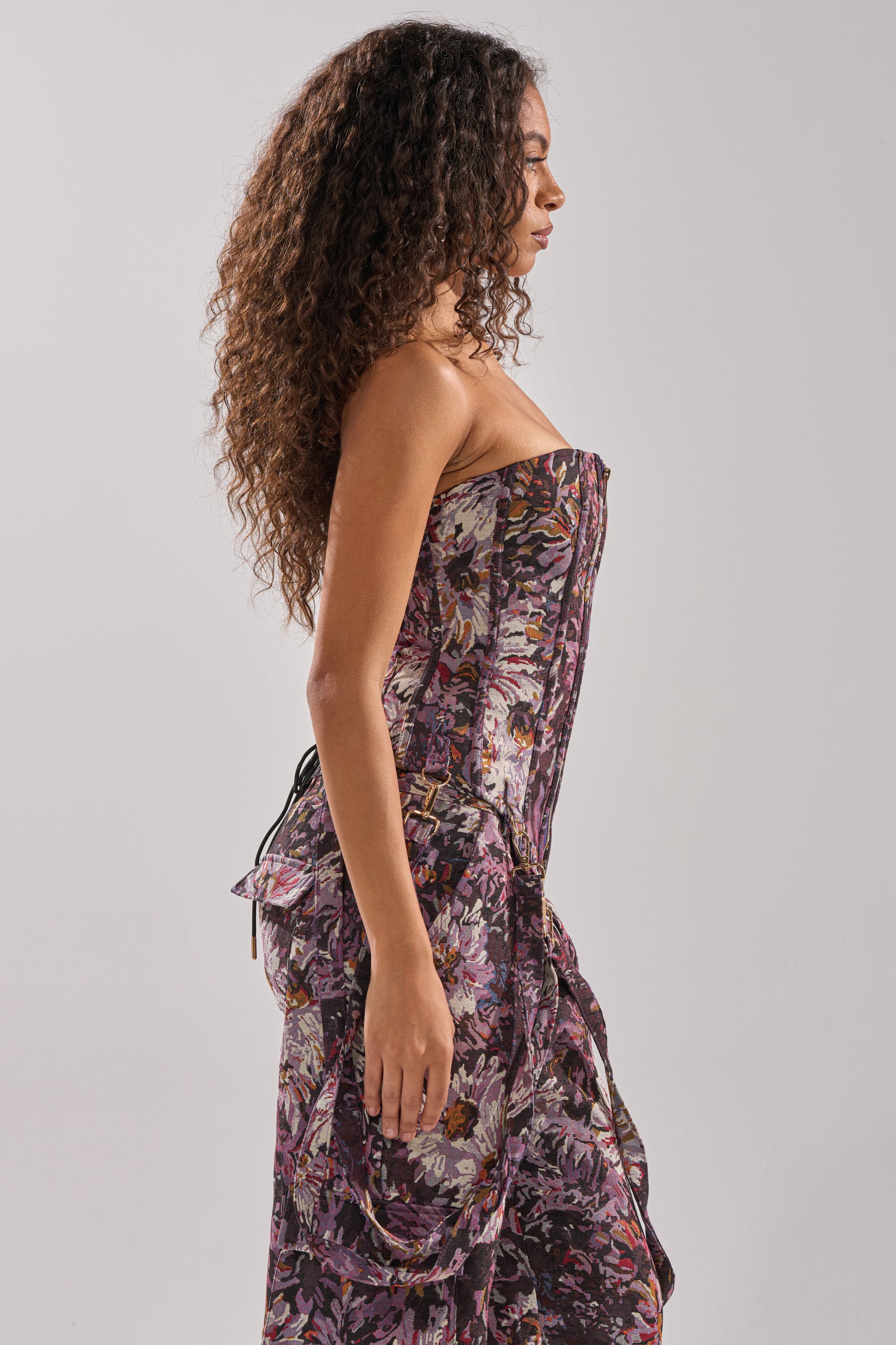 A woman with long curly hair stands in profile, wearing the ABOVE THE MIDDLE FLORAL CORSET, a strapless floral dress with ruffled details, against a plain light gray background.
