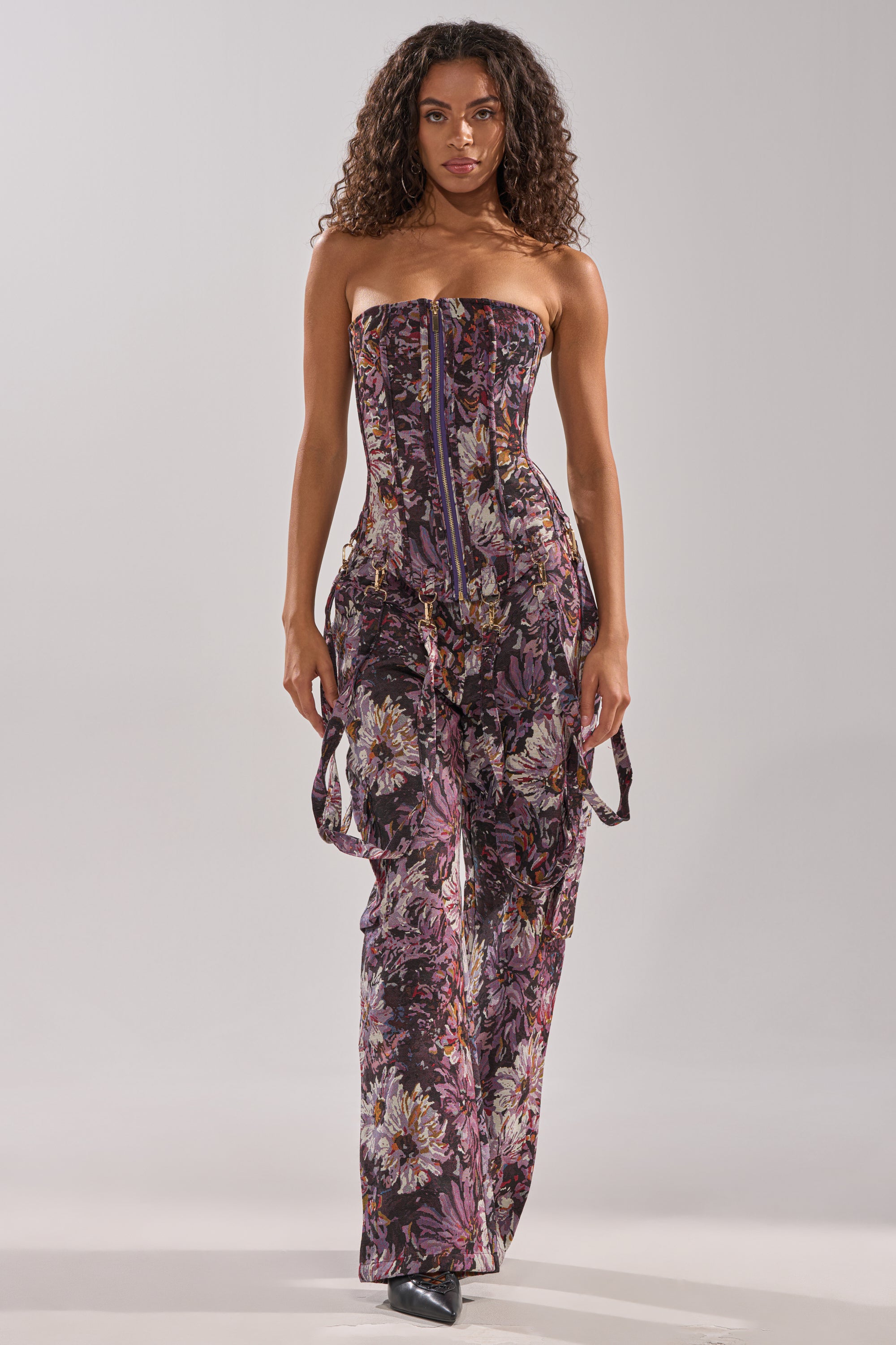 A curly-haired woman confidently models the ABOVE THE MIDDLE FLORAL CORSET, a strapless jumpsuit with flared pants in purple, pink, and black floral print, against a plain light background.