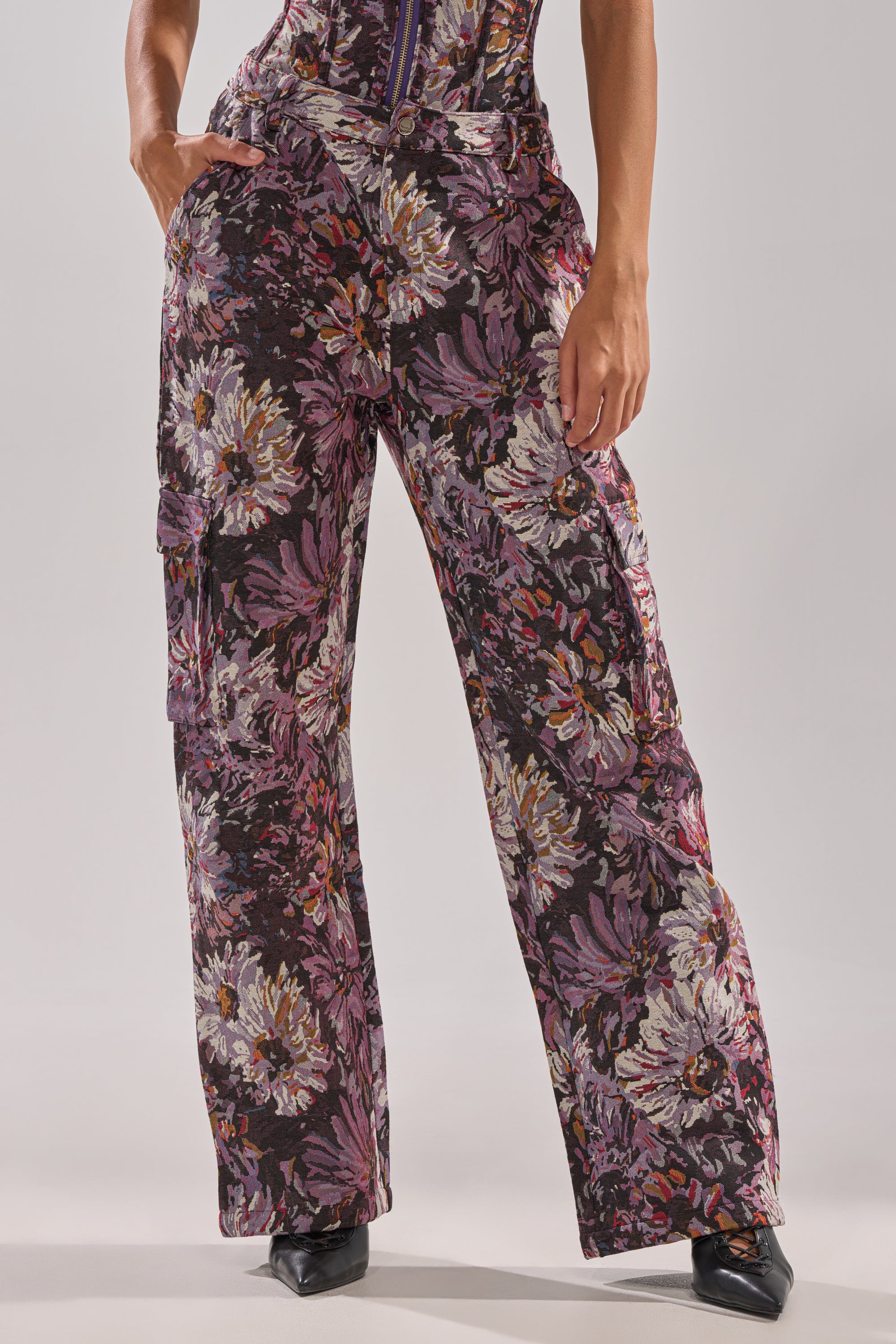 A person wears the ABOVE THE MIDDLE FLORAL TROUSER—high-rise, wide-leg trousers in vibrant purple, white, and green floral print—with black pointed-toe shoes. One hand is casually tucked into a pocket, highlighting the relaxed silhouette.