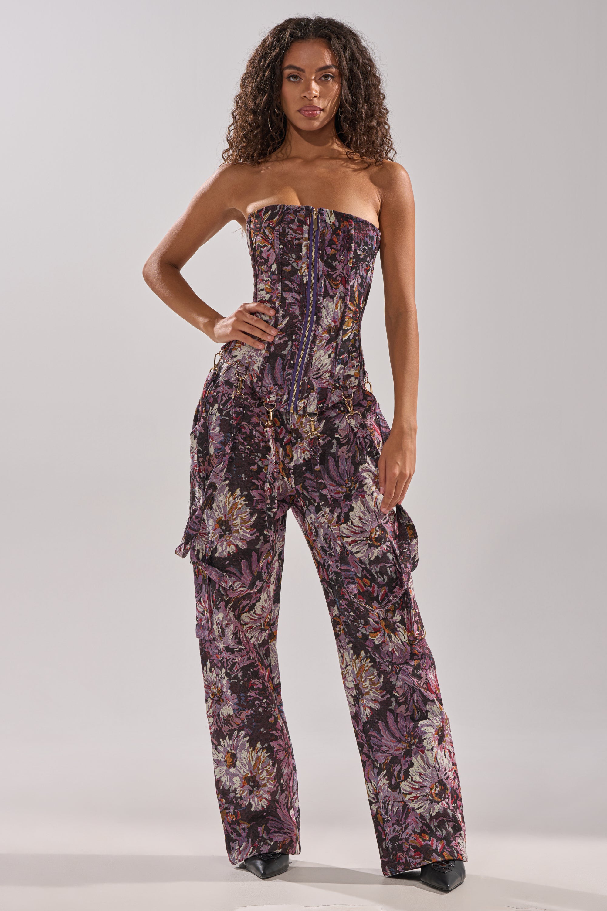 A woman with curly hair models the ABOVE THE MIDDLE FLORAL TROUSER, a wide-leg, strapless jumpsuit with a floral pattern in purple, pink, and black. She stands confidently with one hand on her hip against a plain backdrop.
