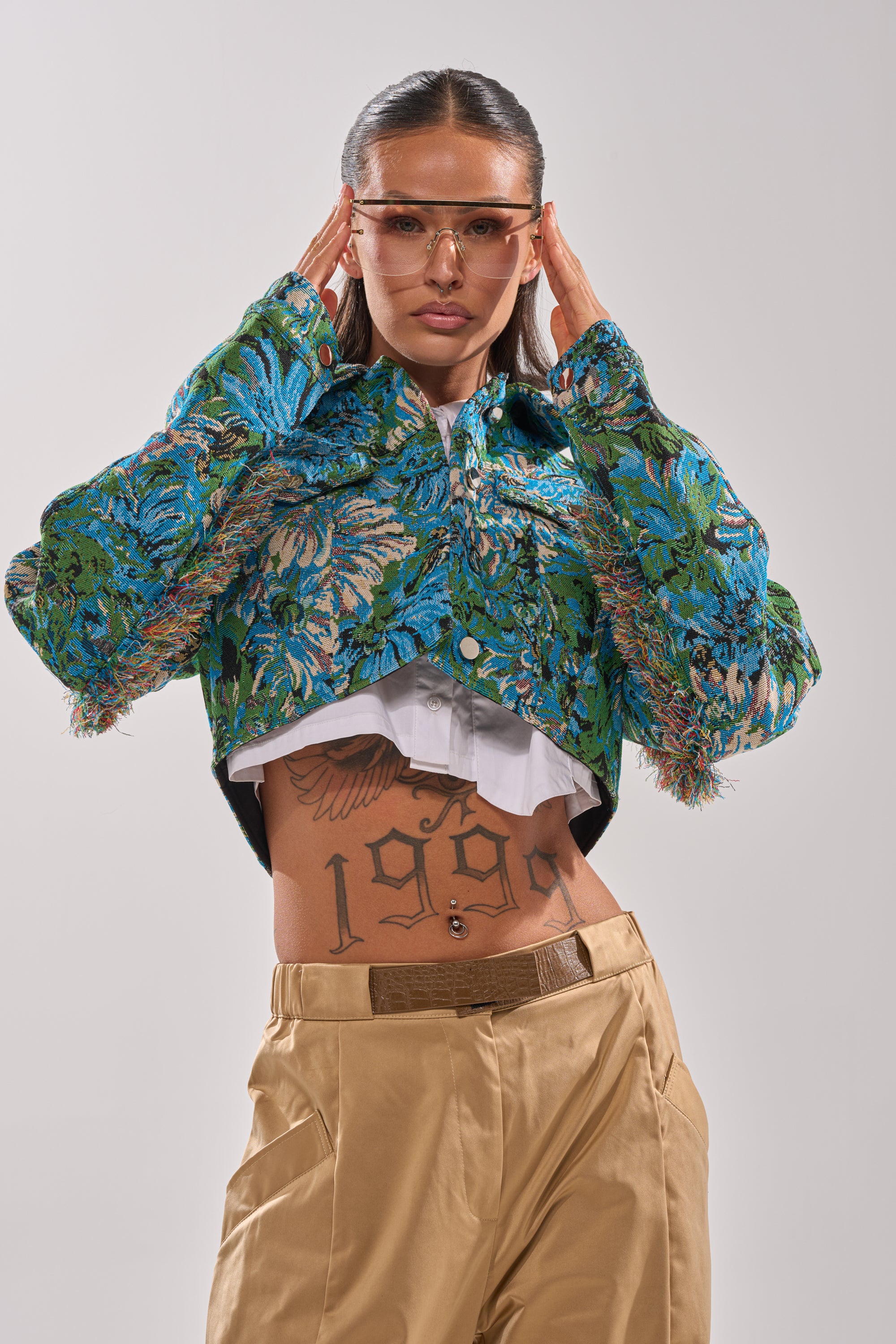 A person wearing tan pants, a white shirt, the TRY ME TAPESTRY BOMBER in green and blue floral jacquard, and orange-tinted glasses poses with hands on their head. Their midriff is exposed, revealing a "1990" tattoo. 0