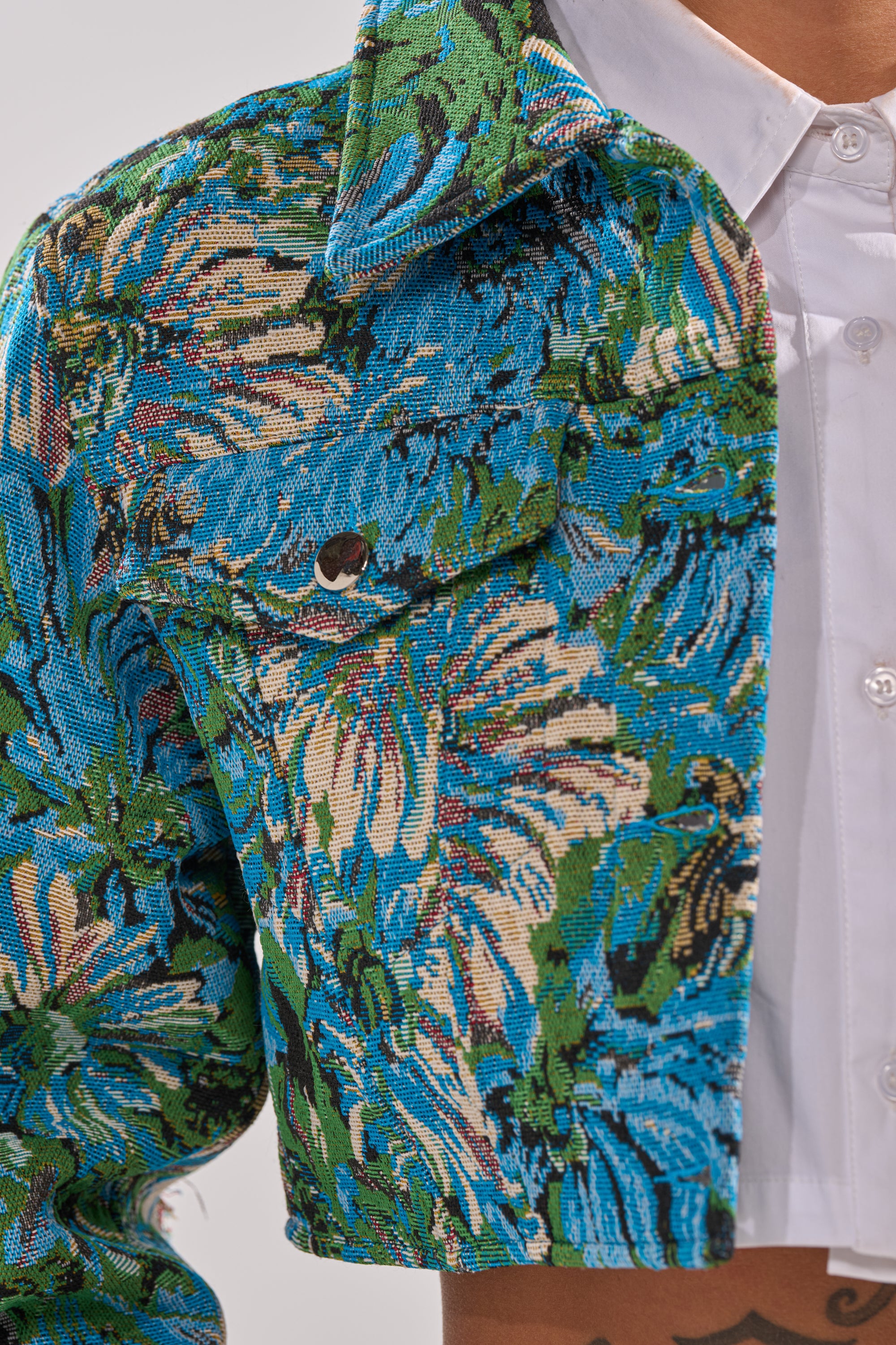 A close-up of a person wearing the TRY ME TAPESTRY BOMBER—featuring a blue, green, and beige floral tapestry fabric with a chest pocket—over a white collared shirt. The photo highlights the textured material and pocket detail. 10