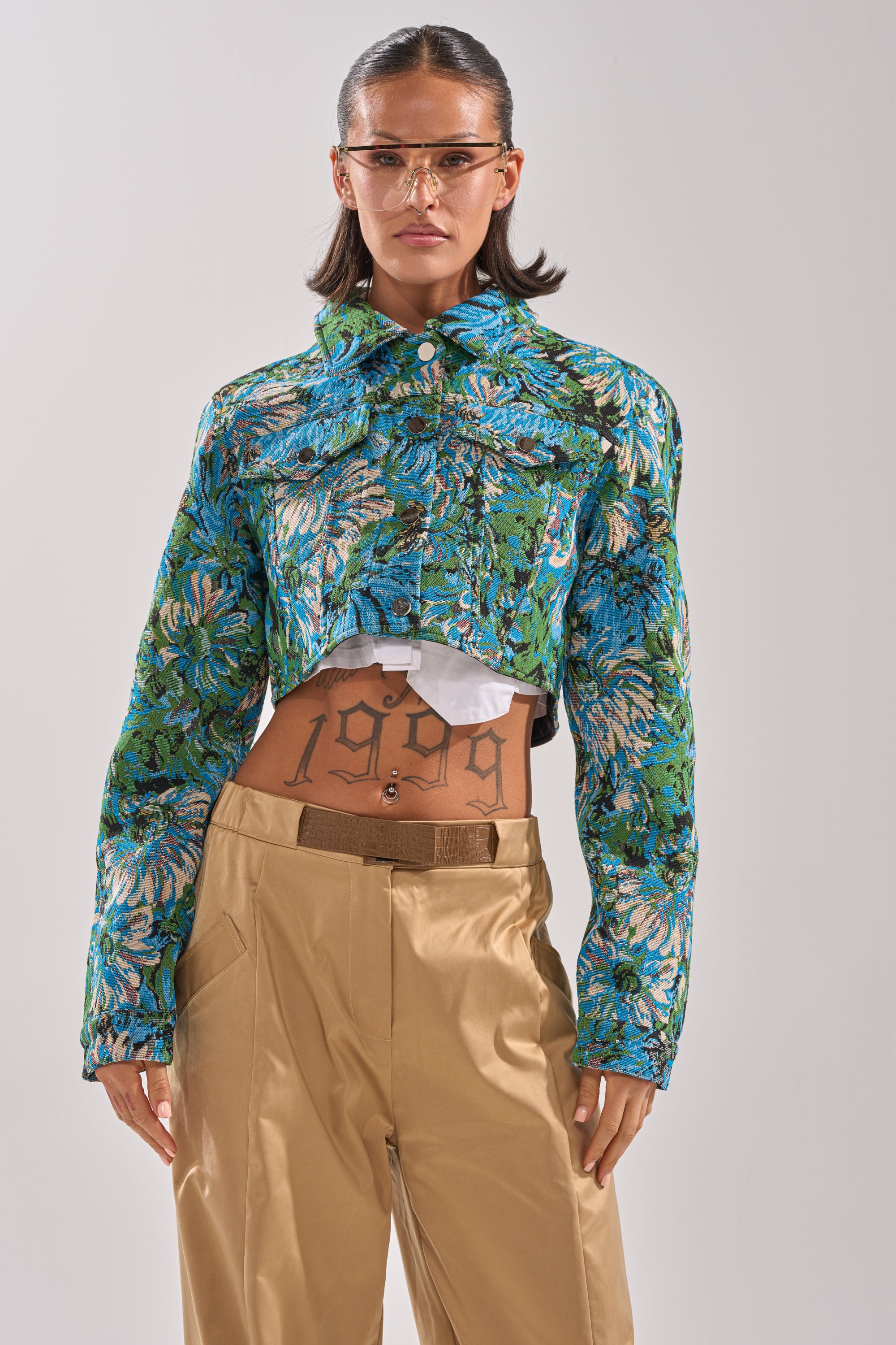 Someone with slicked-back hair and clear glasses wears the TRY ME TAPESTRY BOMBER, featuring blue and green florals, over a white top and beige high-waisted pants. A "1999" tattoo is visible on their stomach. 1