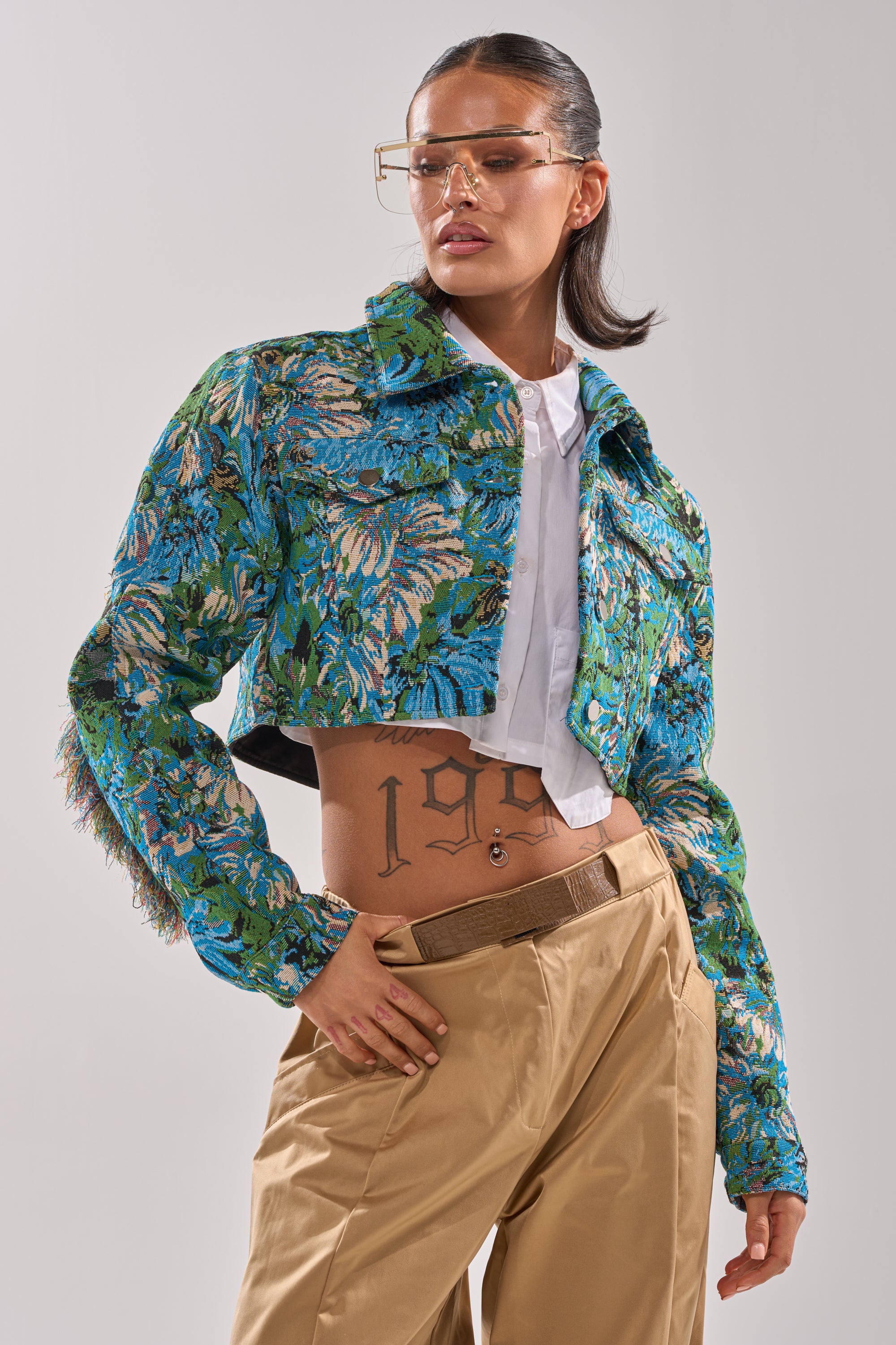 A stylish person poses confidently in the TRY ME TAPESTRY BOMBER, a green and blue floral jacquard jacket, paired with a white shirt, tan pants, clear glasses, hoop earrings, and a "199" tattoo visible across their midriff. 2