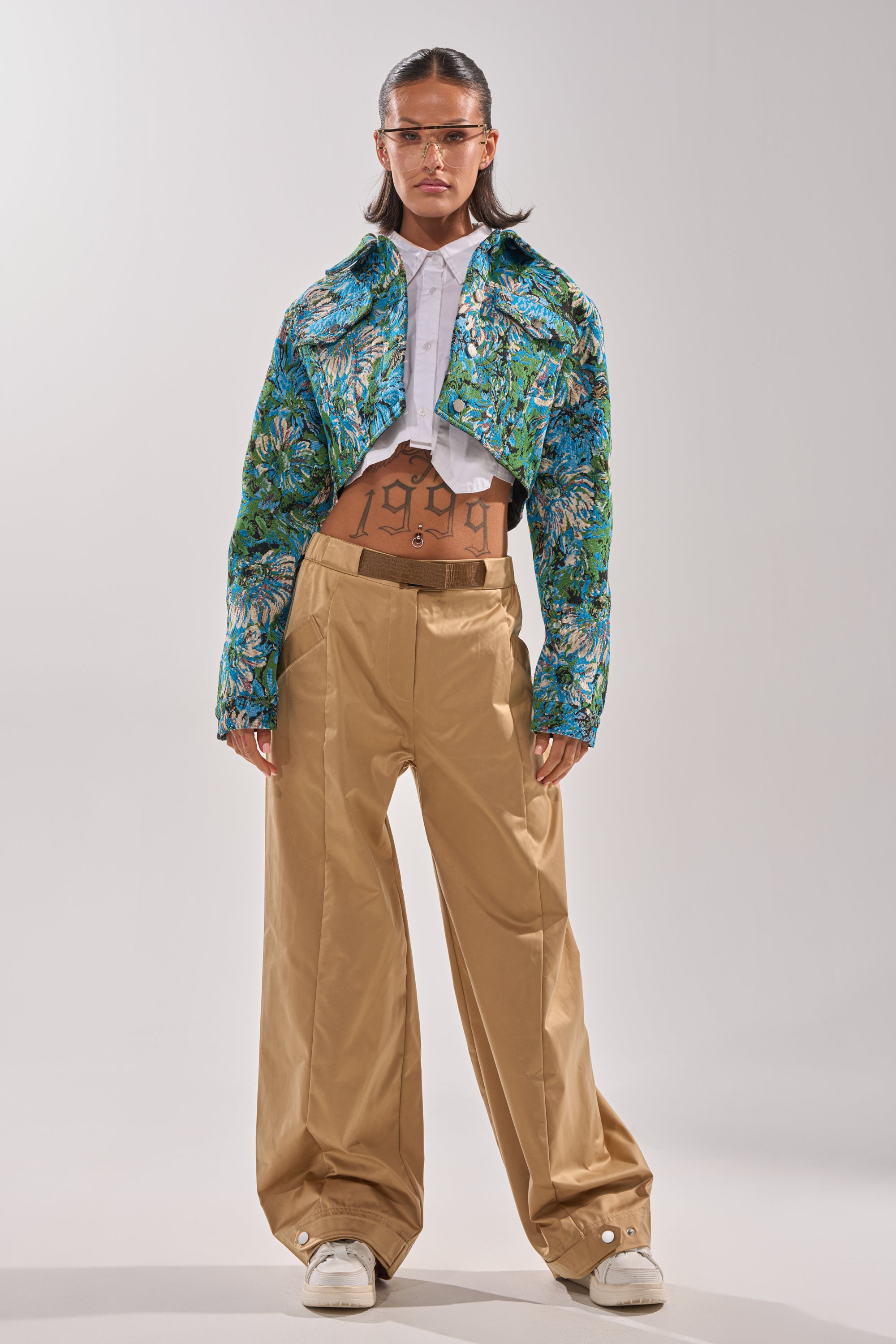 A woman stands facing forward in tan wide-leg pants, a white crop top, and the vibrant TRY ME TAPESTRY BOMBER. She wears clear visor sunglasses, white sneakers, and "1999" is marked on her stomach. 6