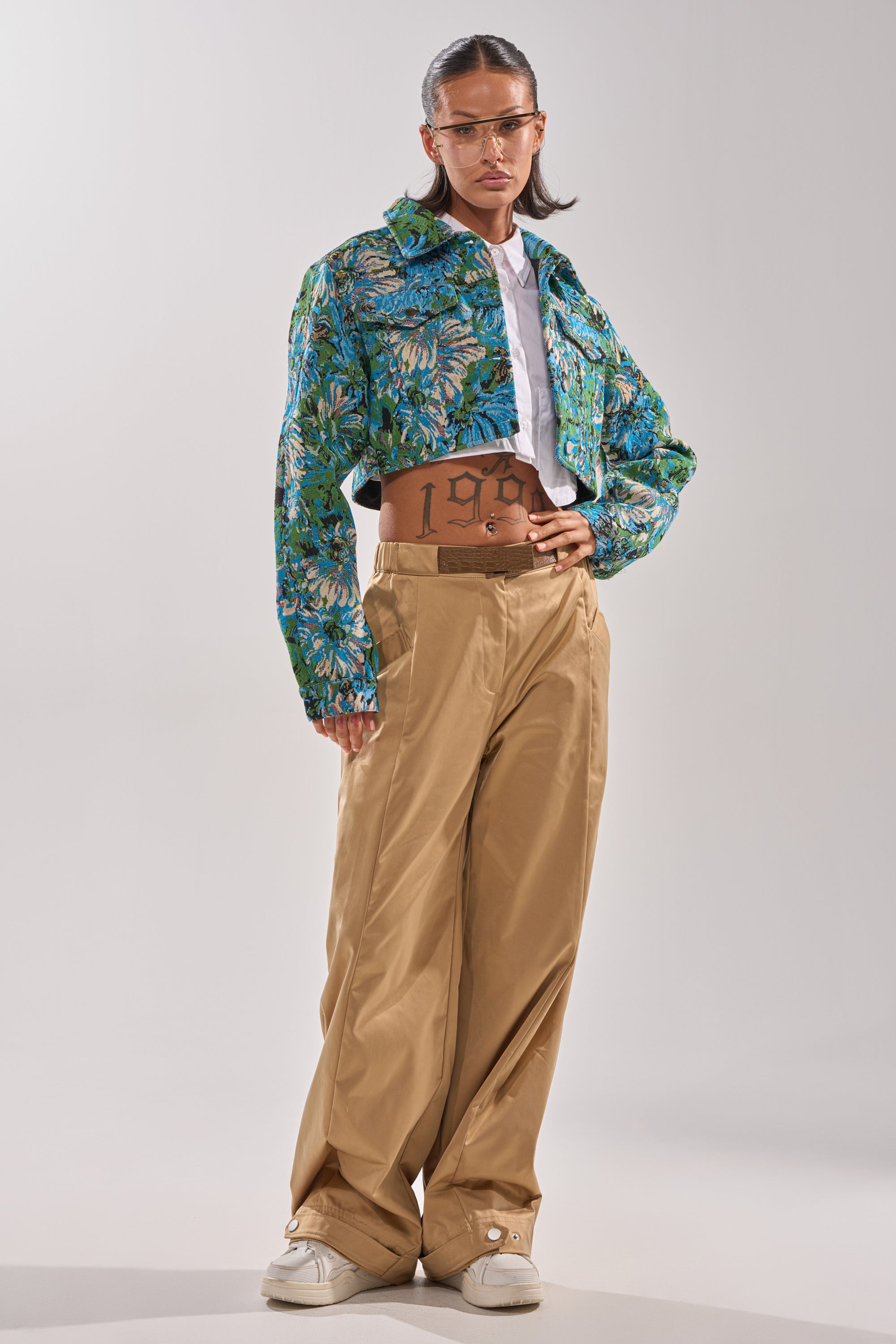 A person stands confidently in the TRY ME TAPESTRY BOMBER, a blue and green floral jacquard cropped jacket, paired with a white shirt, tan wide-leg pants, white sneakers, clear sunglasses, and their hair pulled back tightly. 8
