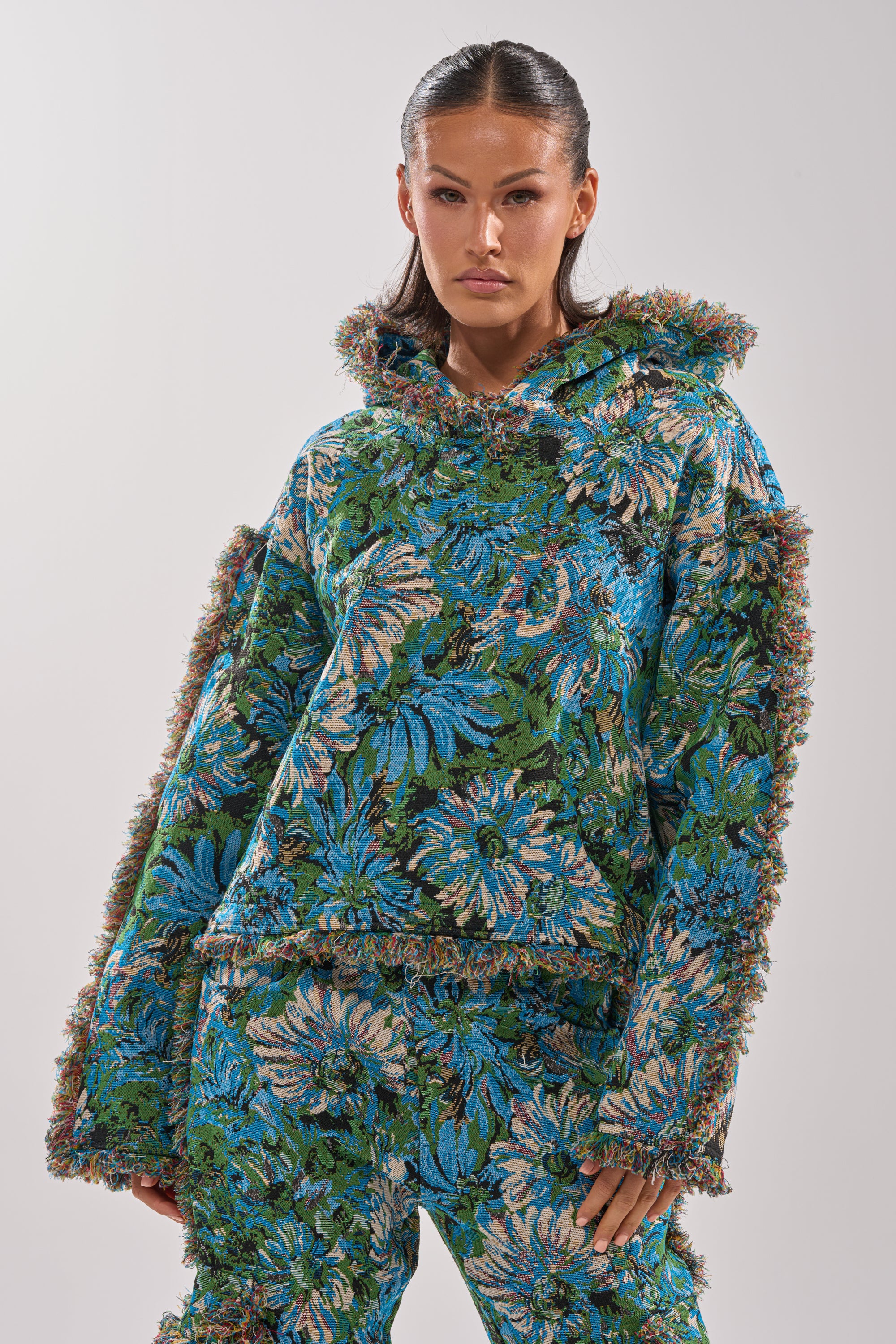 A woman stands confidently in the TRY ME TAPESTRY SWEATSHIRT, a blue and green floral print pullover with textured trim and a hood, paired with loose-fitting pants against a plain light background.