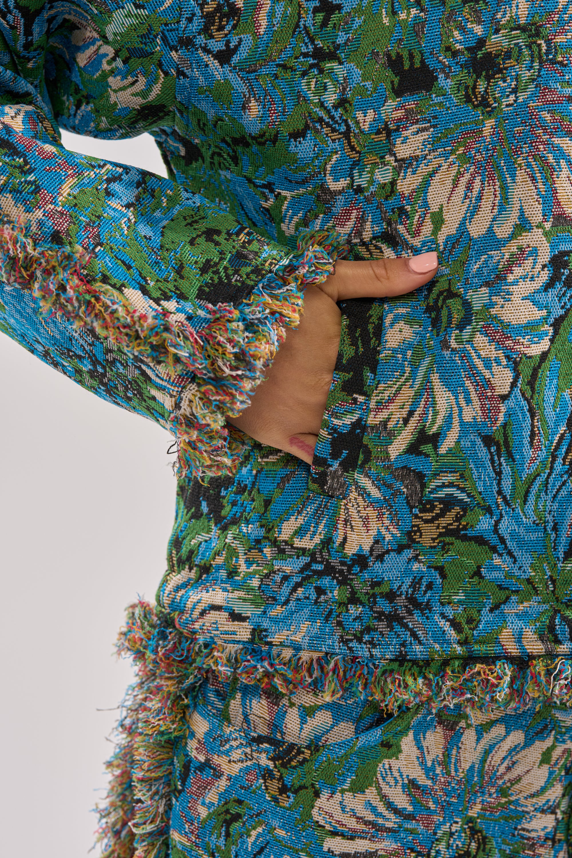 Someone wears the TRY ME TAPESTRY SWEATSHIRT, a vintage-inspired top with a colorful floral pattern in green, blue, and cream. Frayed edges at the sleeves and waist add charm as they slip their hand into a pocket.