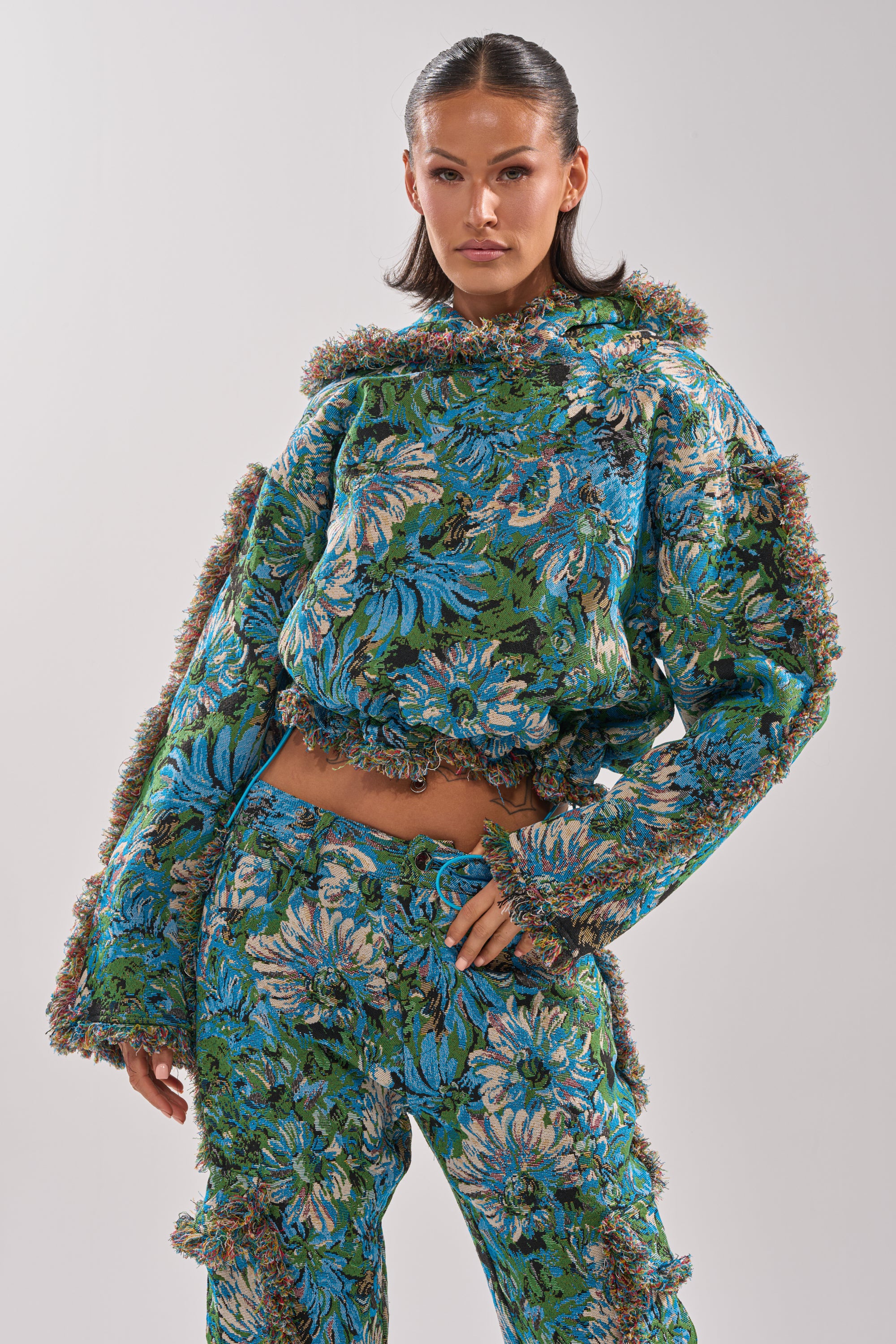 A woman poses confidently in the TRY ME TAPESTRY SWEATSHIRT, featuring a vintage-inspired floral print in blue, green, and beige. The loose-fit sweatshirt is paired with matching fringed pants, creating a bold and colorful look against a plain background.