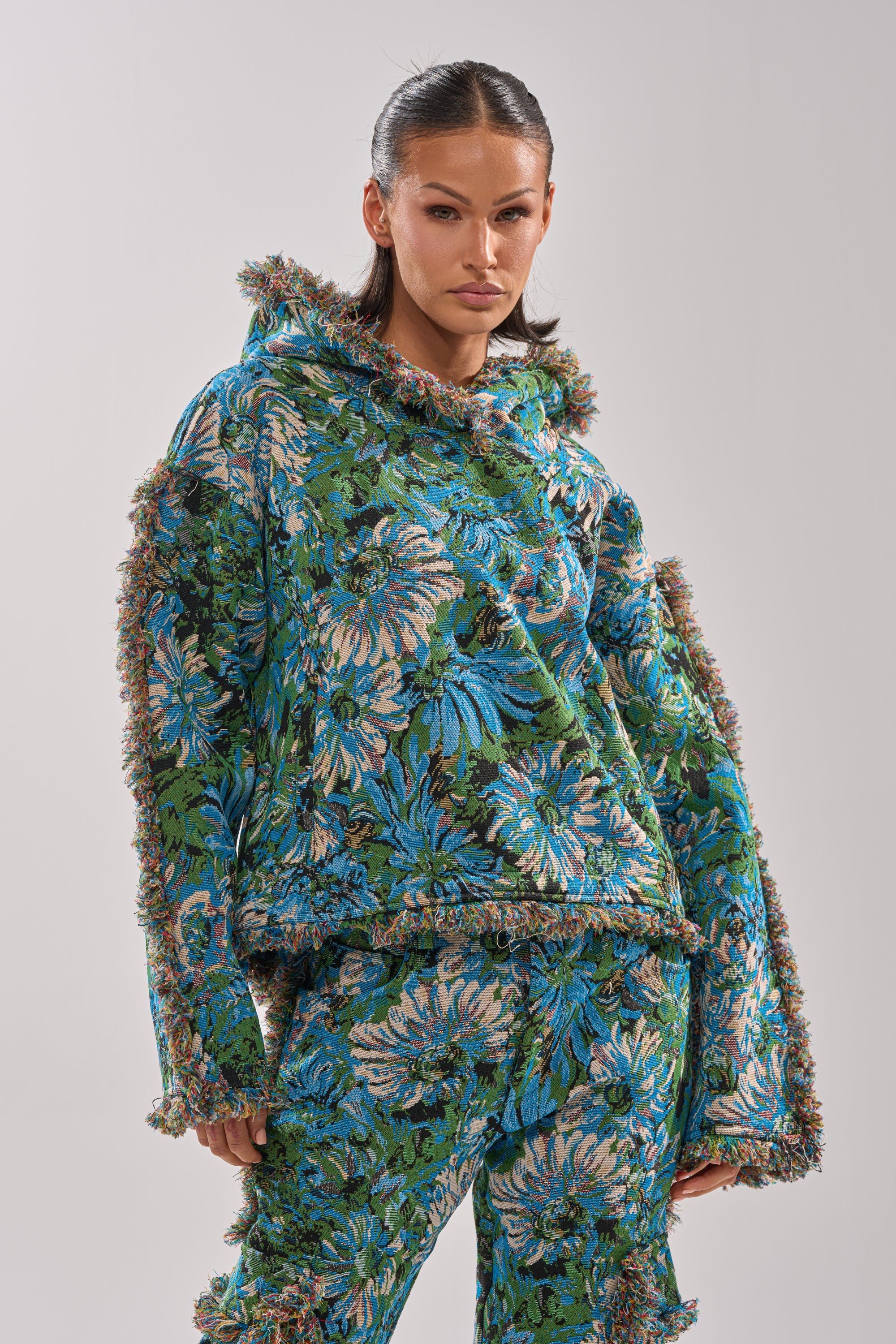 Wearing the TRY ME TAPESTRY SWEATSHIRT featuring blue, green, and beige floral patterns with fringe edges, a person stands against a plain light gray background, highlighting vintage-inspired style.