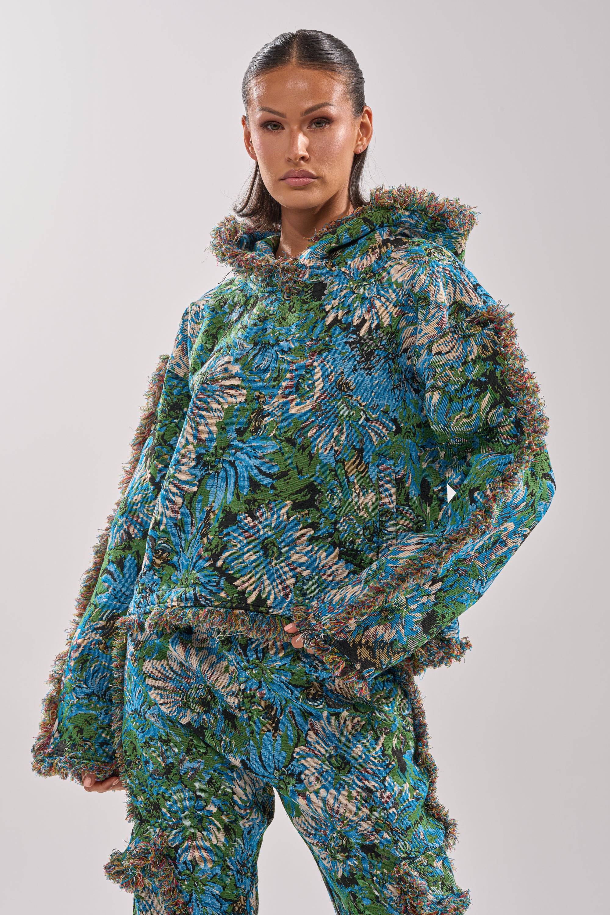 A woman poses confidently in the TRY ME TAPESTRY SWEATSHIRT and matching pants, featuring bold blue, green, and brown florals with textured, fringed edges against a plain light background.
