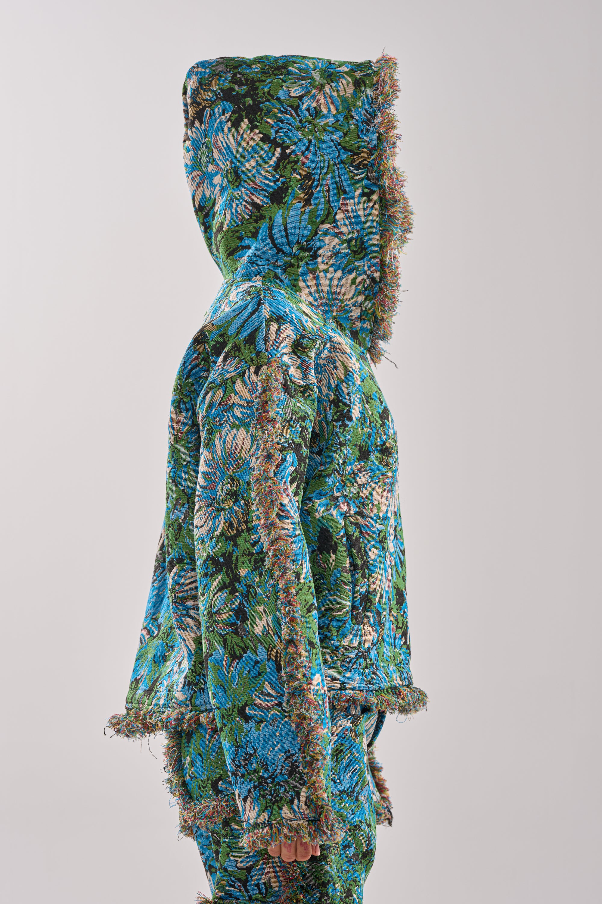 Someone in profile wears the TRY ME TAPESTRY SWEATSHIRT, featuring a textured blue and green floral pattern with frayed edges, channeling vintage-inspired artistry against a plain light background.