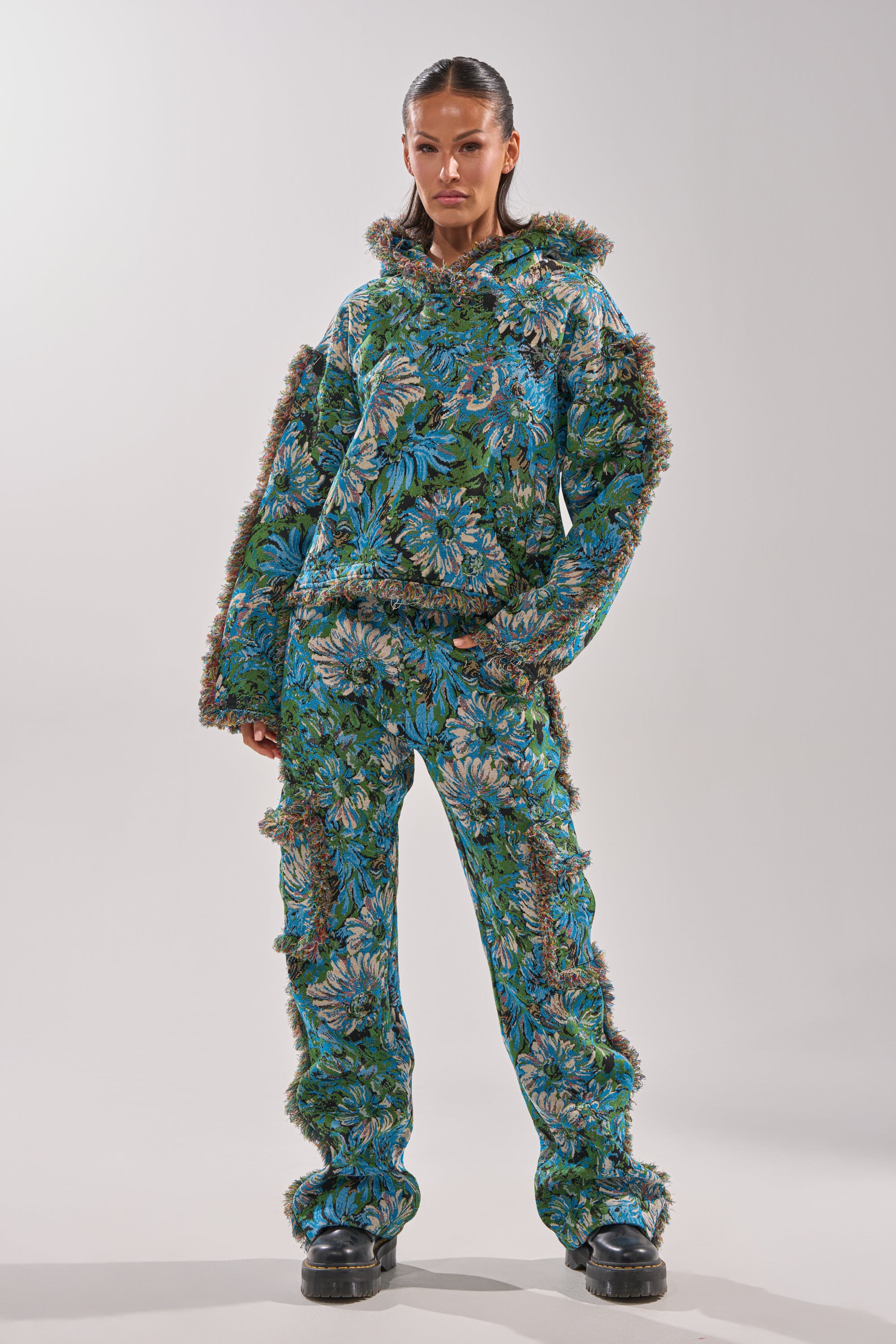 Wearing the TRY ME TAPESTRY SWEATSHIRT, a person poses in a blue-green floral hooded jacket with fuzzy trim, brown accents, and chunky black shoes—a look that channels vintage sweater charm.