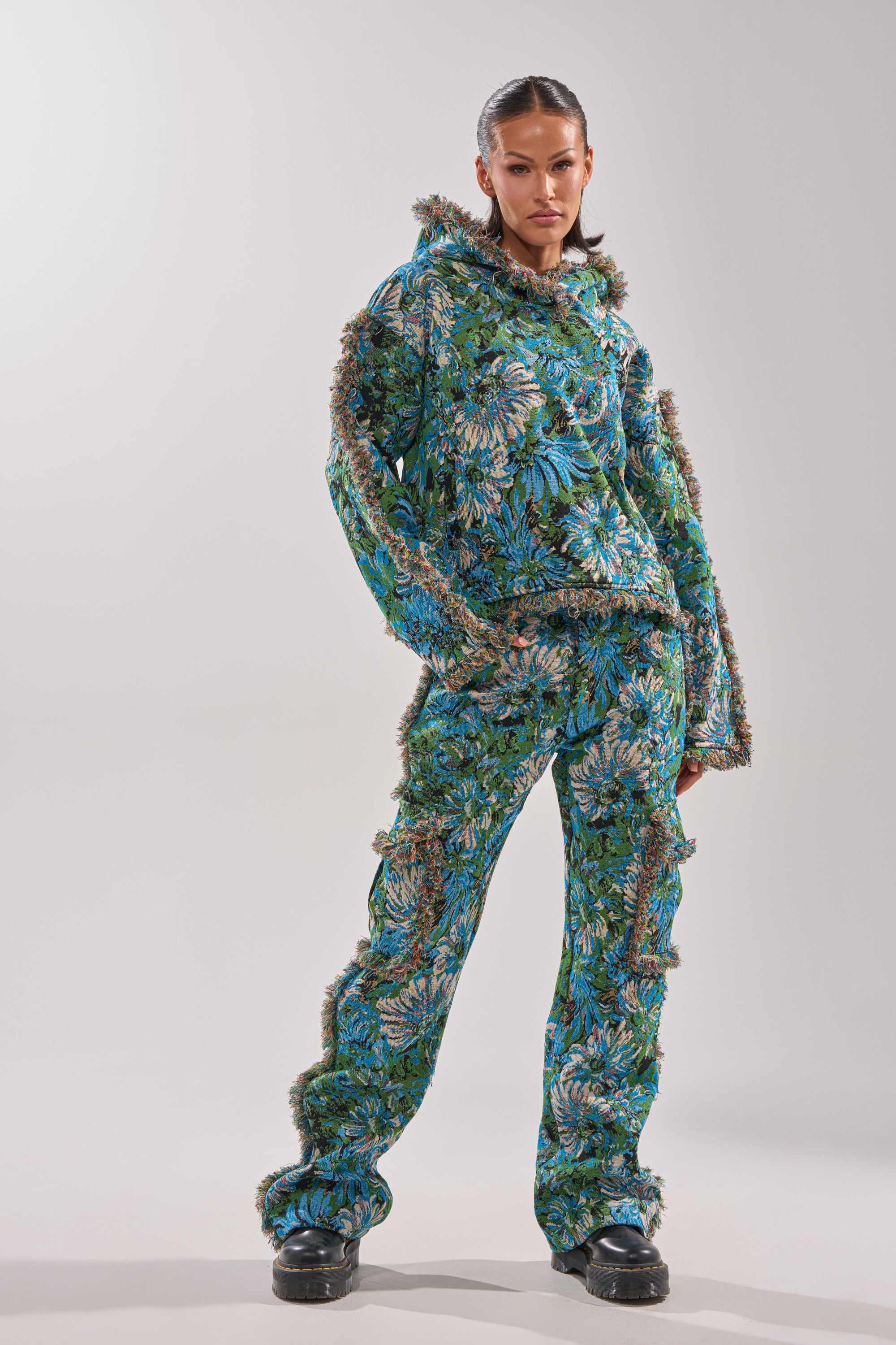 A person stands confidently against a plain light gray background, wearing the TRY ME TAPESTRY SWEATSHIRT with matching pants and fuzzy trim, slicked-back hair, and black platform shoes.