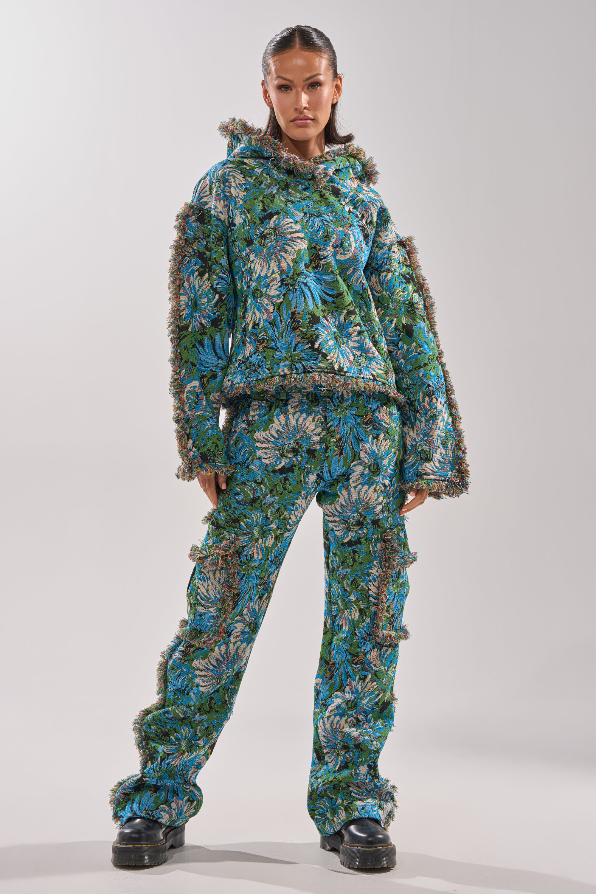 A woman stands confidently in TRY ME TAPESTRY TROUSER—blue and green floral-patterned, jacquard brocade, straight-leg pants with fuzzy edging—paired with a matching hoodie and black chunky shoes against a light gray background.