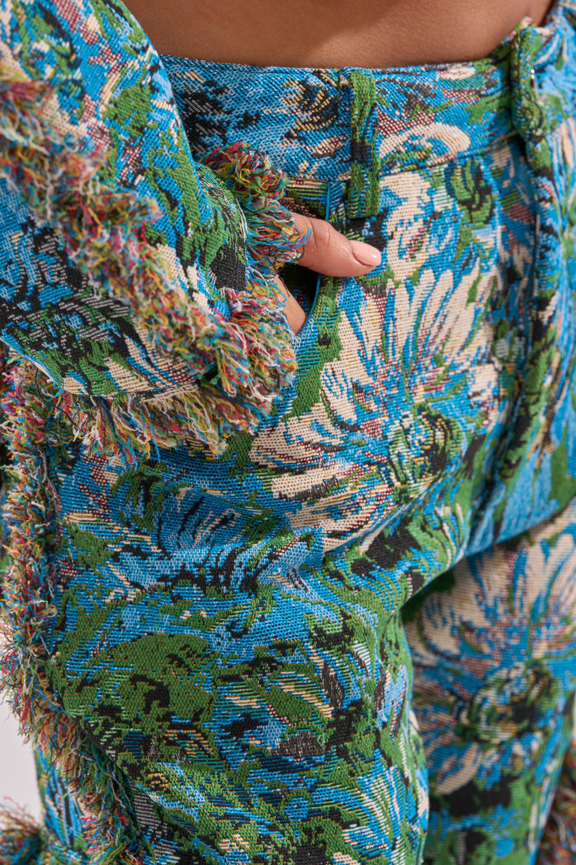 Close-up of a person wearing the TRY ME TAPESTRY TROUSER and matching jacket, featuring a colorful blue, green, and beige floral jacquard brocade pattern. Their hand with painted nails is tucked into the trouser pocket.