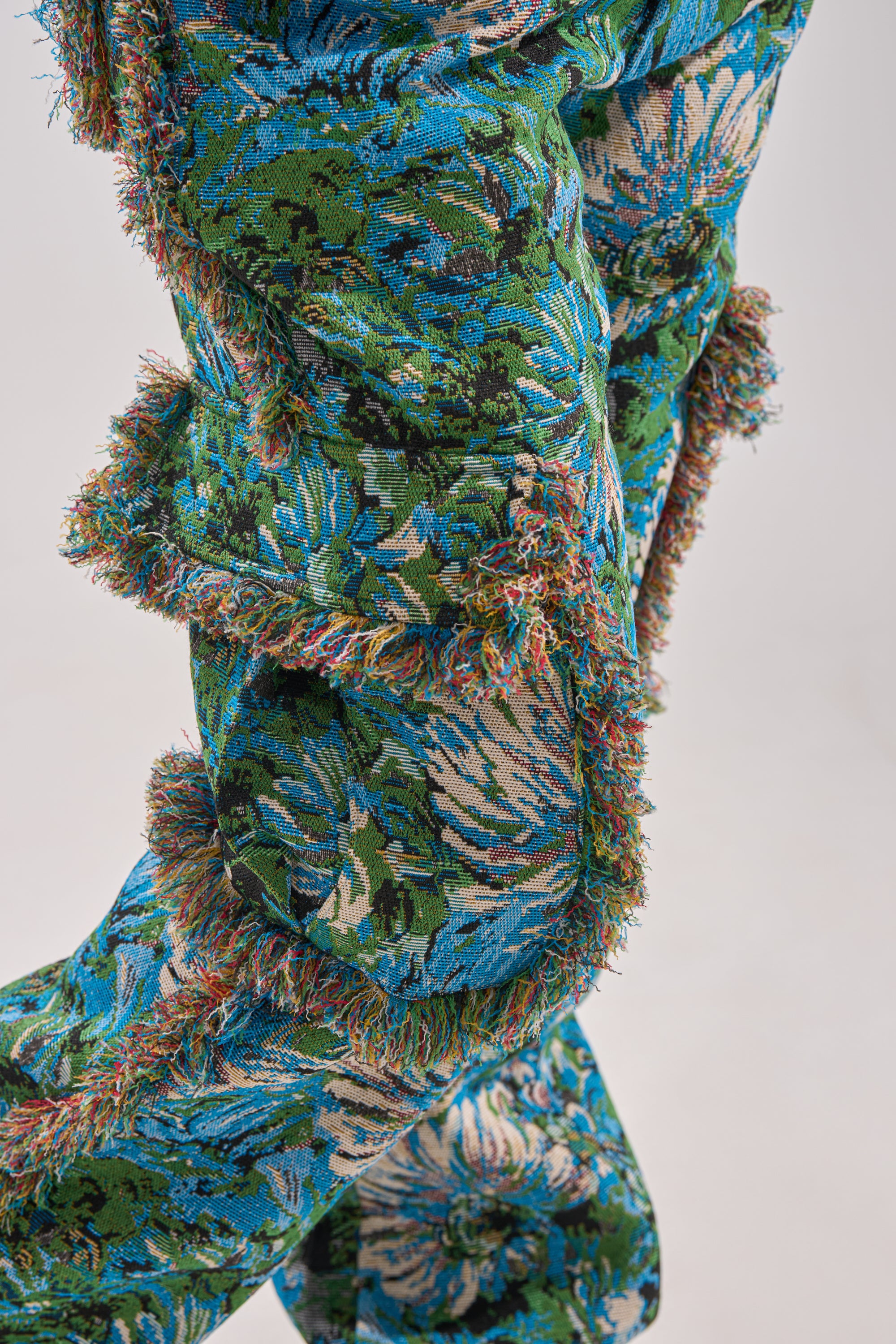 Close-up of the TRY ME TAPESTRY TROUSER: jacquard brocade pants with blue, green, and beige floral and leaf patterns, textured fabric, fringed edges, and visible seams joining each section.