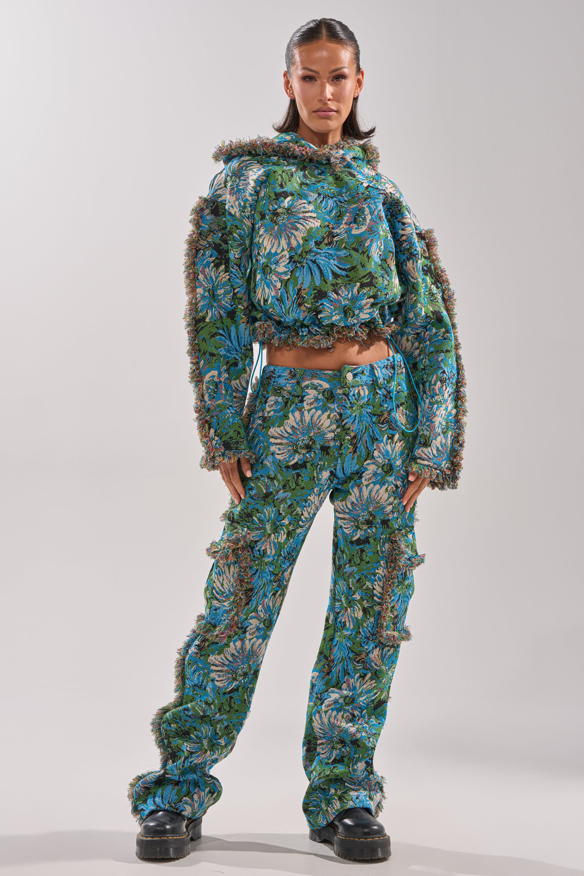 A woman stands confidently in the TRY ME TAPESTRY TROUSER, a jacquard brocade pant with textured fringed edges, paired with a matching cropped floral top and chunky black shoes, her hair sleek and styled.