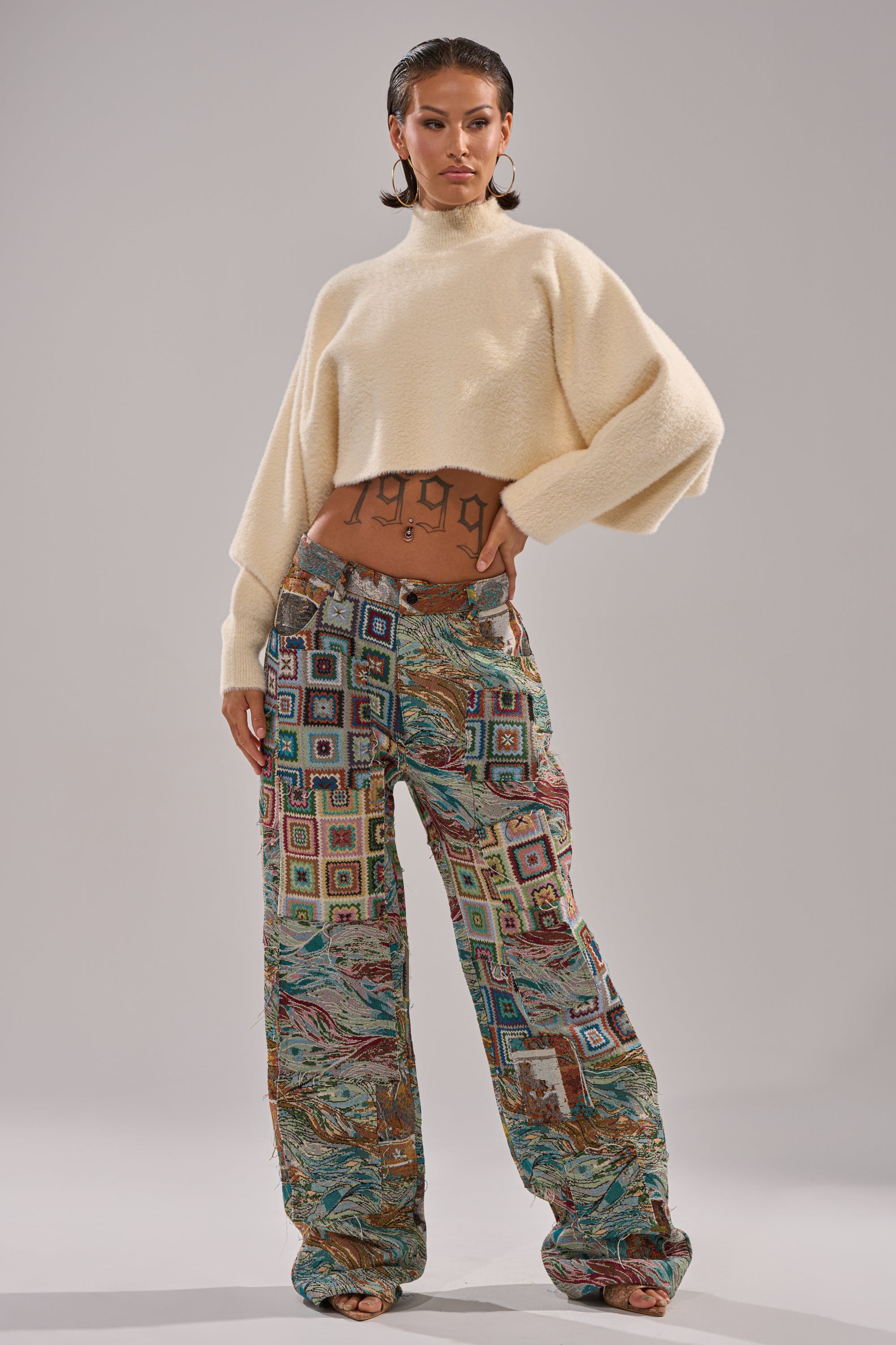 A confident individual wears high-waisted WILD PATCHWORK TROUSER and a cream cropped sweater with wide sleeves, showing geometric tattoos on their midriff, set against a plain light background.